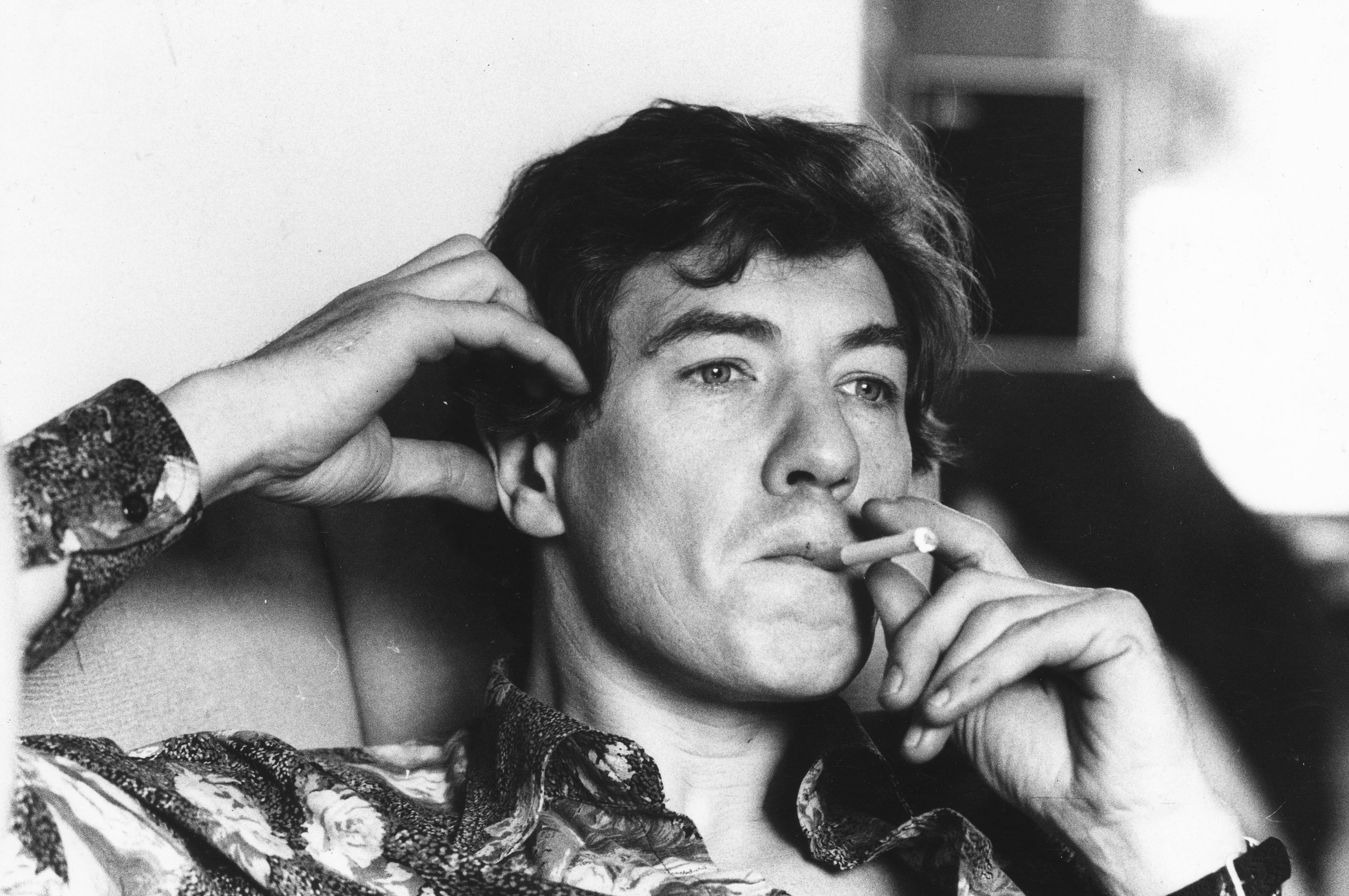 Ian McKellen is seen smoking a cigarette, sitting contemplatively with one hand behind his head. He is wearing a patterned shirt
