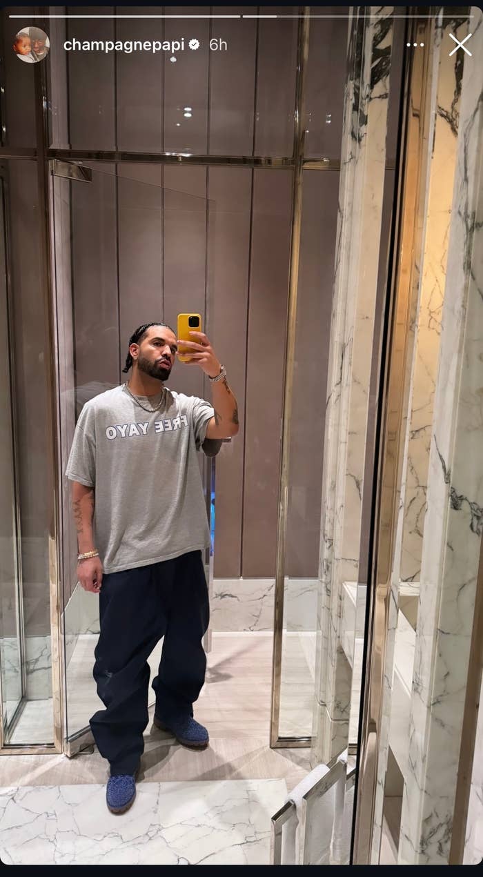 Drake takes a mirror selfie wearing a casual outfit: a grey shirt with &quot;FREE YAYO&quot; printed on it, dark pants, and blue shoes