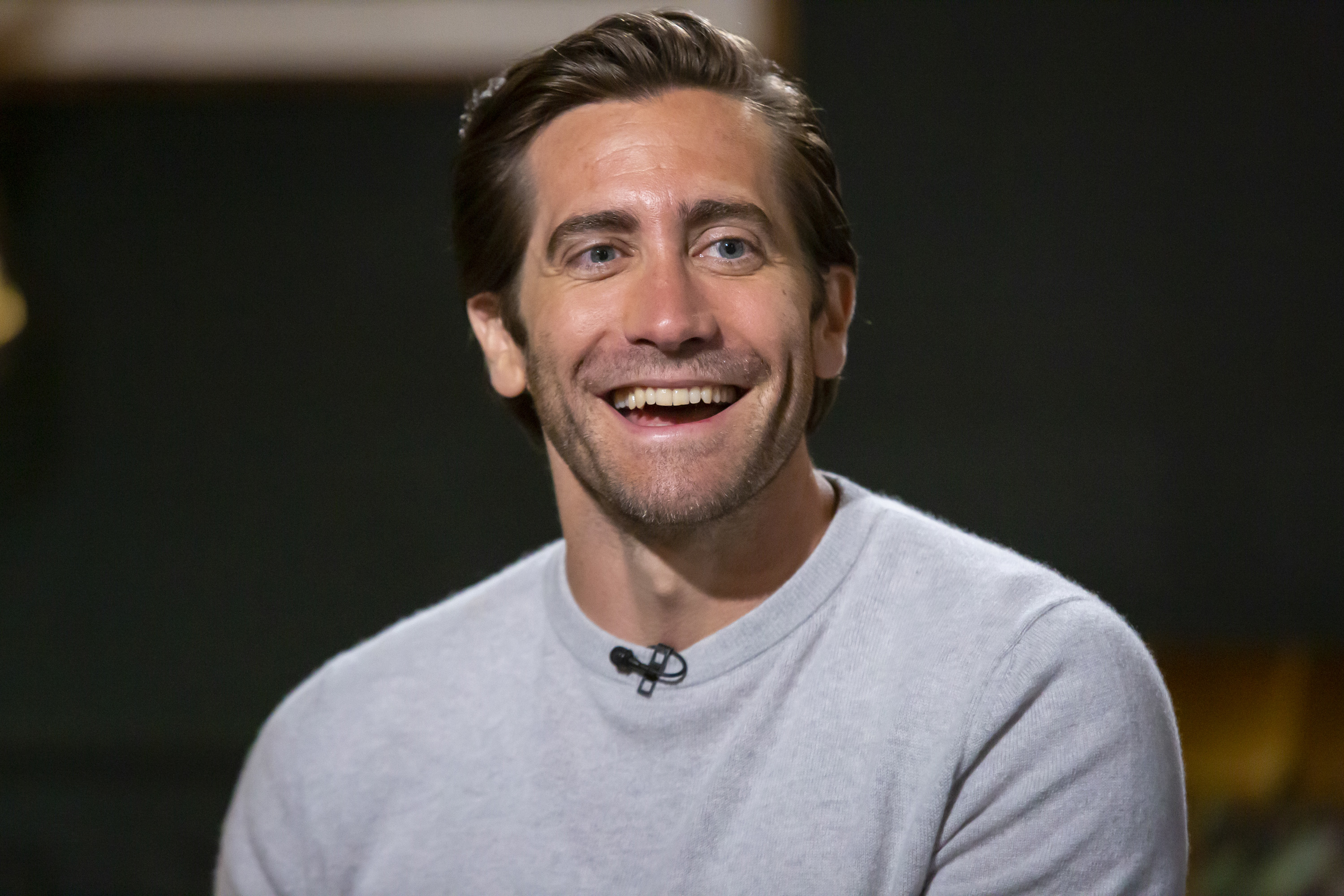 Jake Gyllenhaal smiles while seated, wearing a light-colored sweater, during an interview