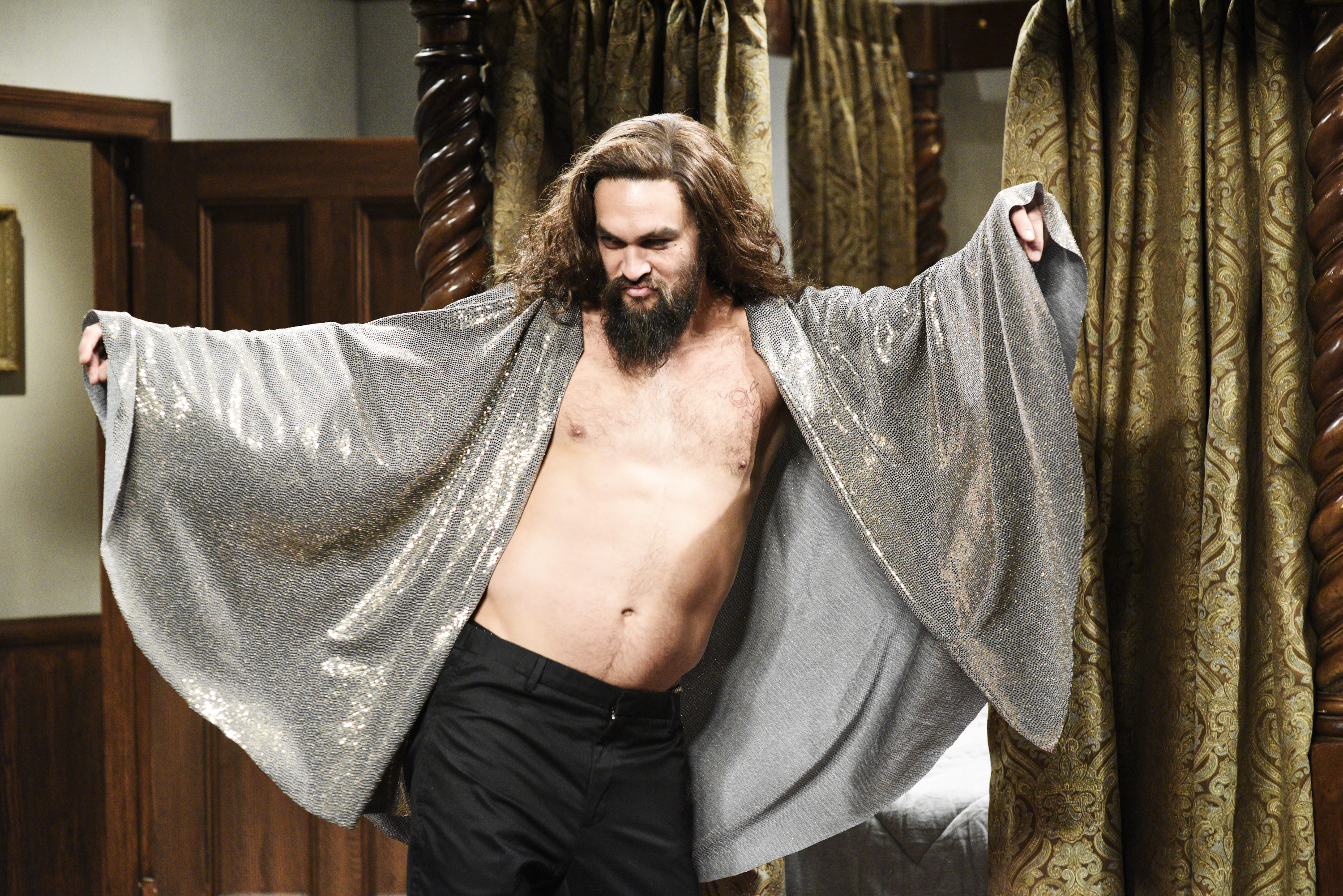 Jason Momoa humorously posing with arms outstretched, wearing a shiny, open, flowing robe, and black pants. The setting appears to be a comedic or theatrical stage