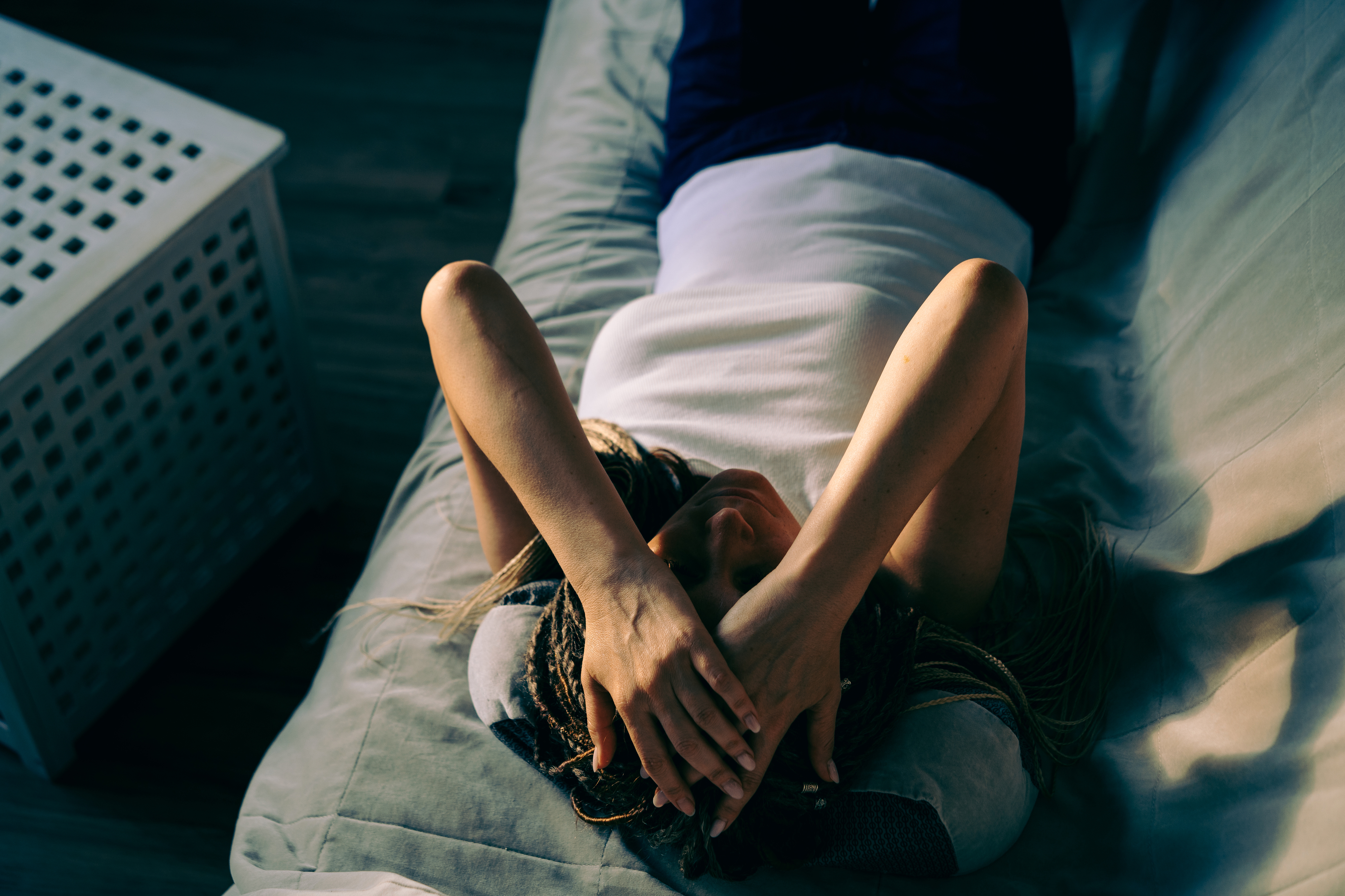 A person is lying on a bed with their arms covering their face, appearing relaxed. The room has a wooden floor and a white laundry basket is visible next to the bed