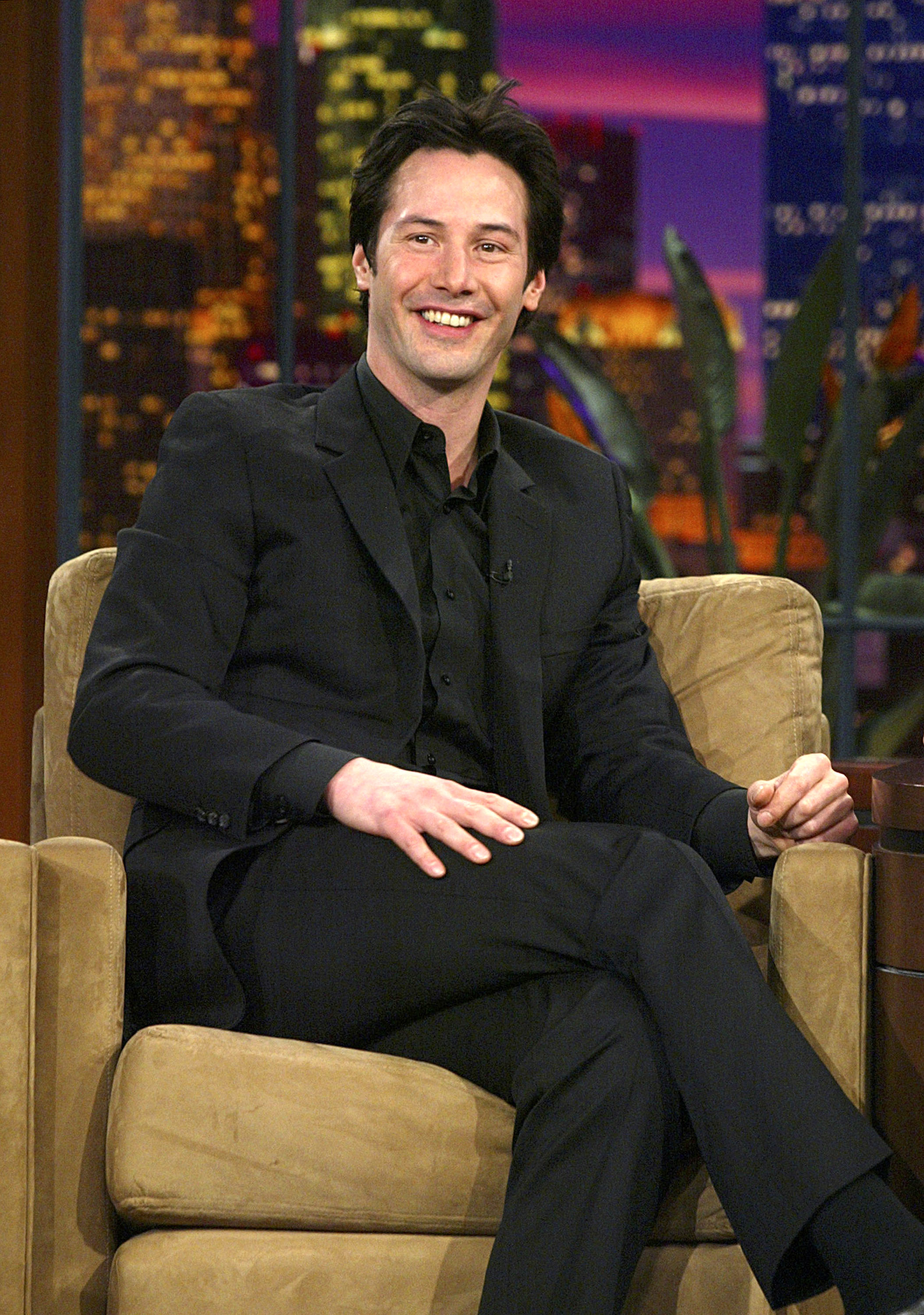 Keanu Reeves is seated on a talk show set, smiling and wearing a black suit