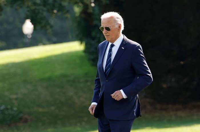 Joe Biden in a navy suit and tie, walking outdoors with sunglasses on