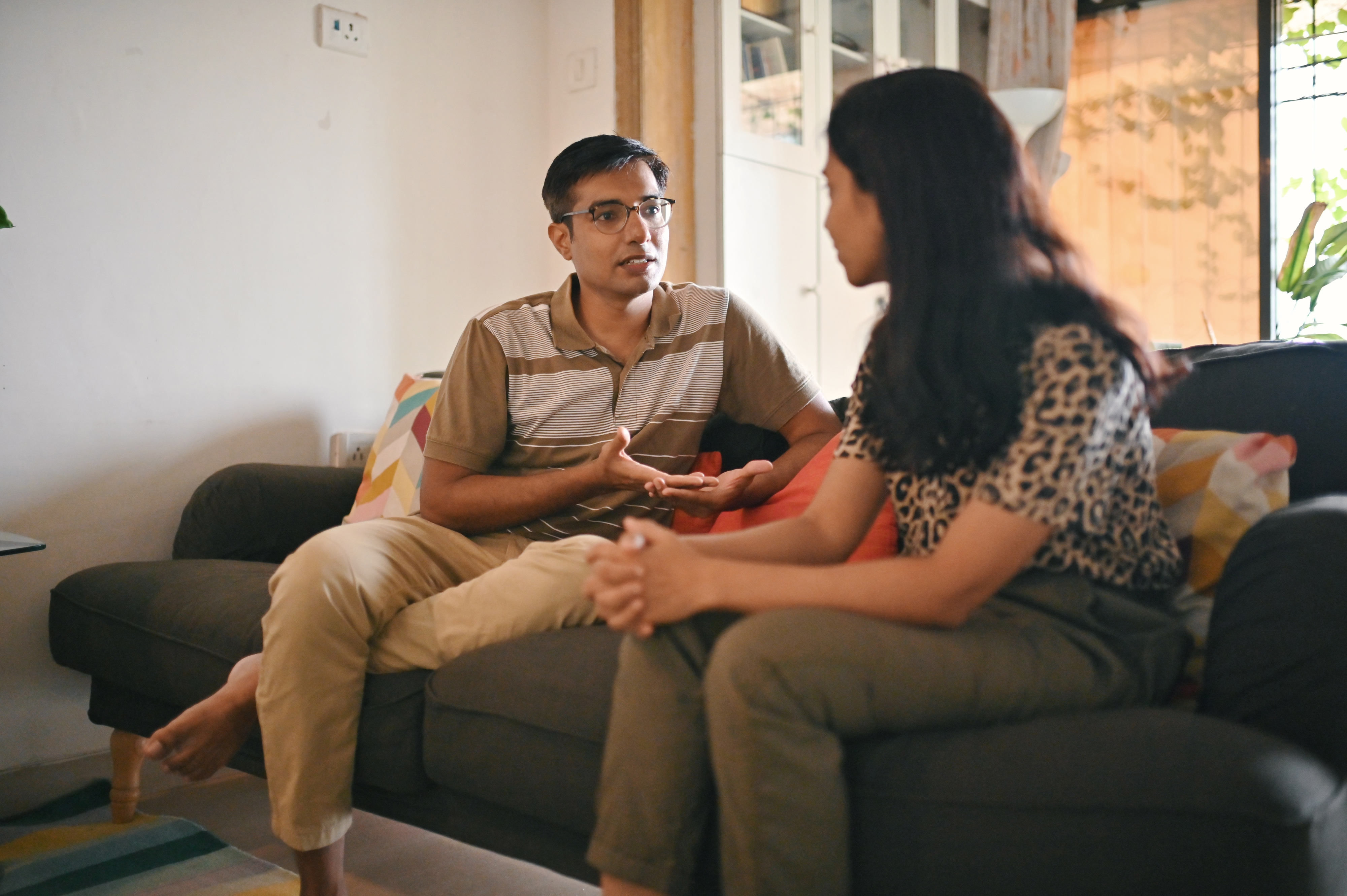 A couple sits on a couch engaged in a serious conversation. The man gestures with his hands while the woman listens attentively