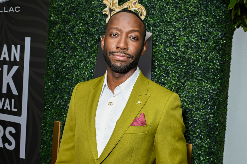 Lamorne Morris in a tailored suit poses at an event with a greenery backdrop