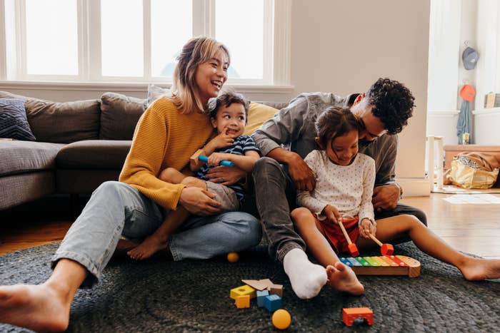 A family of four sits on the floor in a living room, smiling and playing with toy blocks and a xylophone