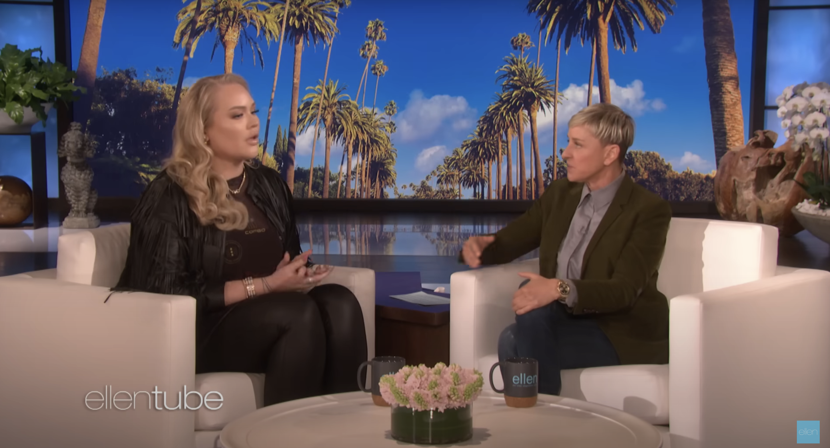 Nikkie de Jager and Ellen DeGeneres sit in white chairs talking on 'The Ellen DeGeneres Show' set, featuring palm trees and blue skies in the background