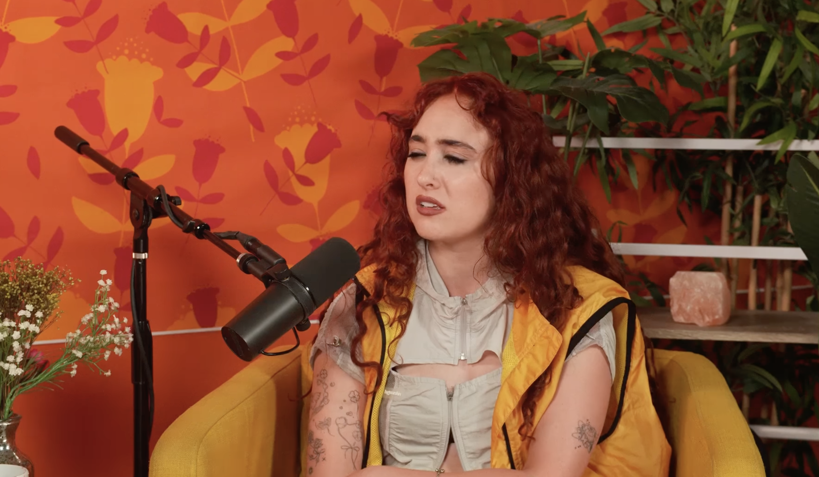 A person with long curly hair sits in a yellow chair speaking into a microphone. They are wearing a light-colored top and a yellow vest