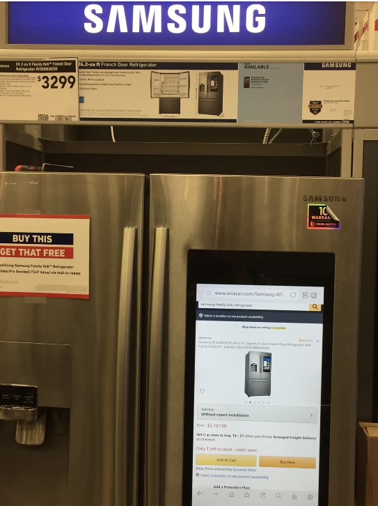 Samsung refrigerator display at a store with a price tag of $3299. The screen on the fridge shows the product page from Amazon