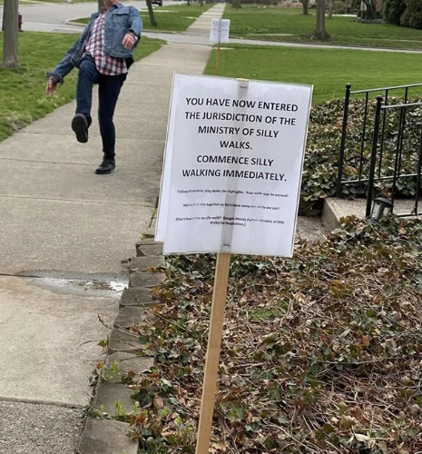 A person walks in a comedic manner on a sidewalk next to a sign that reads, "You have now entered the jurisdiction of the Ministry of Silly Walks. Commence silly walking immediately."