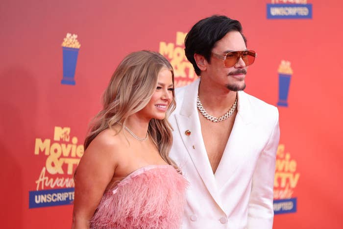 Ariana Madix and Tom Sandoval at the MTV Movie & TV Awards. Ariana wears a feathered strapless dress, and Tom wears a white suit with a chain necklace and sunglasses