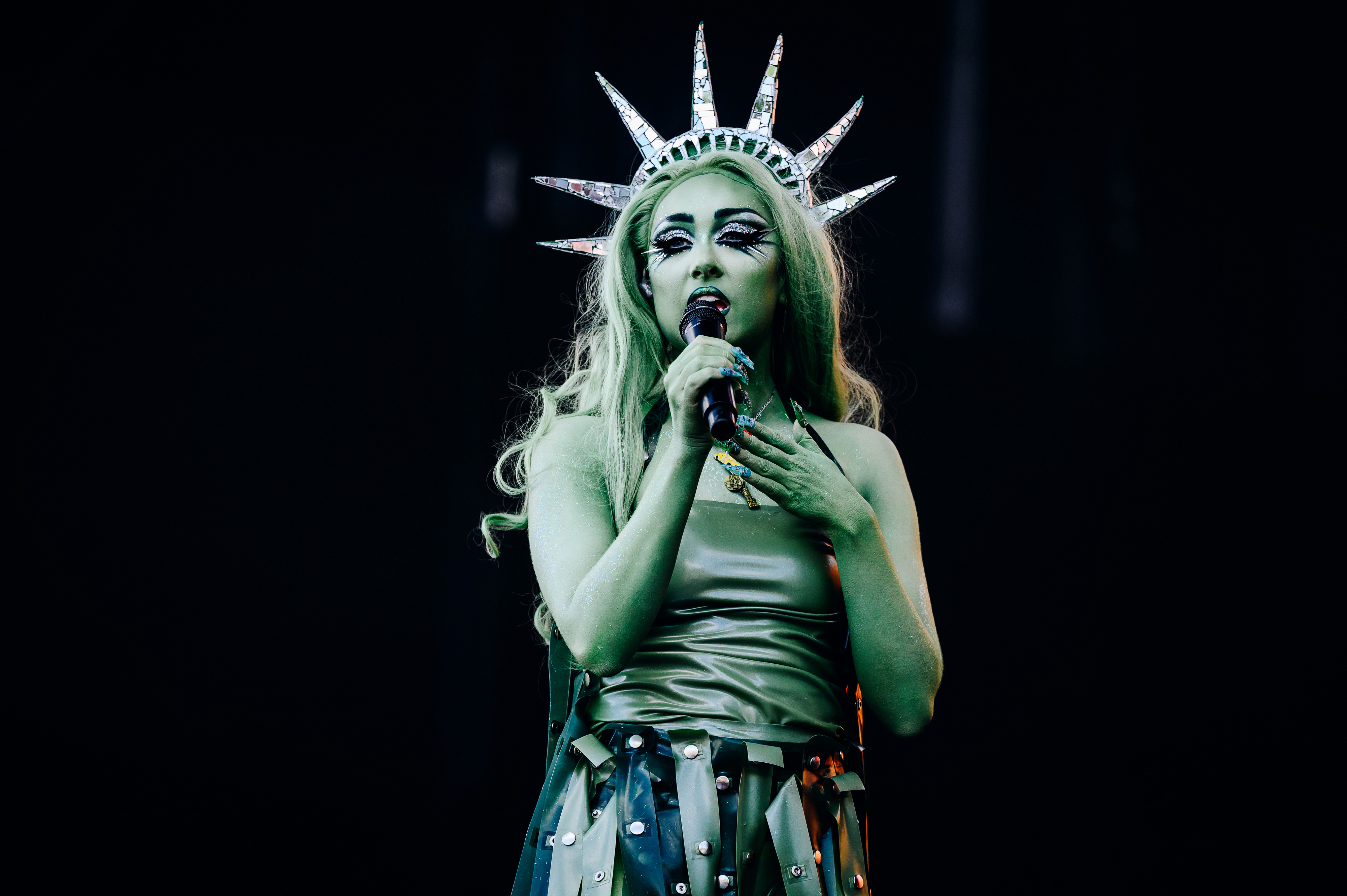 Person dressed as the Statue of Liberty sings into a microphone, wearing a crown with spikes and a metallic outfit