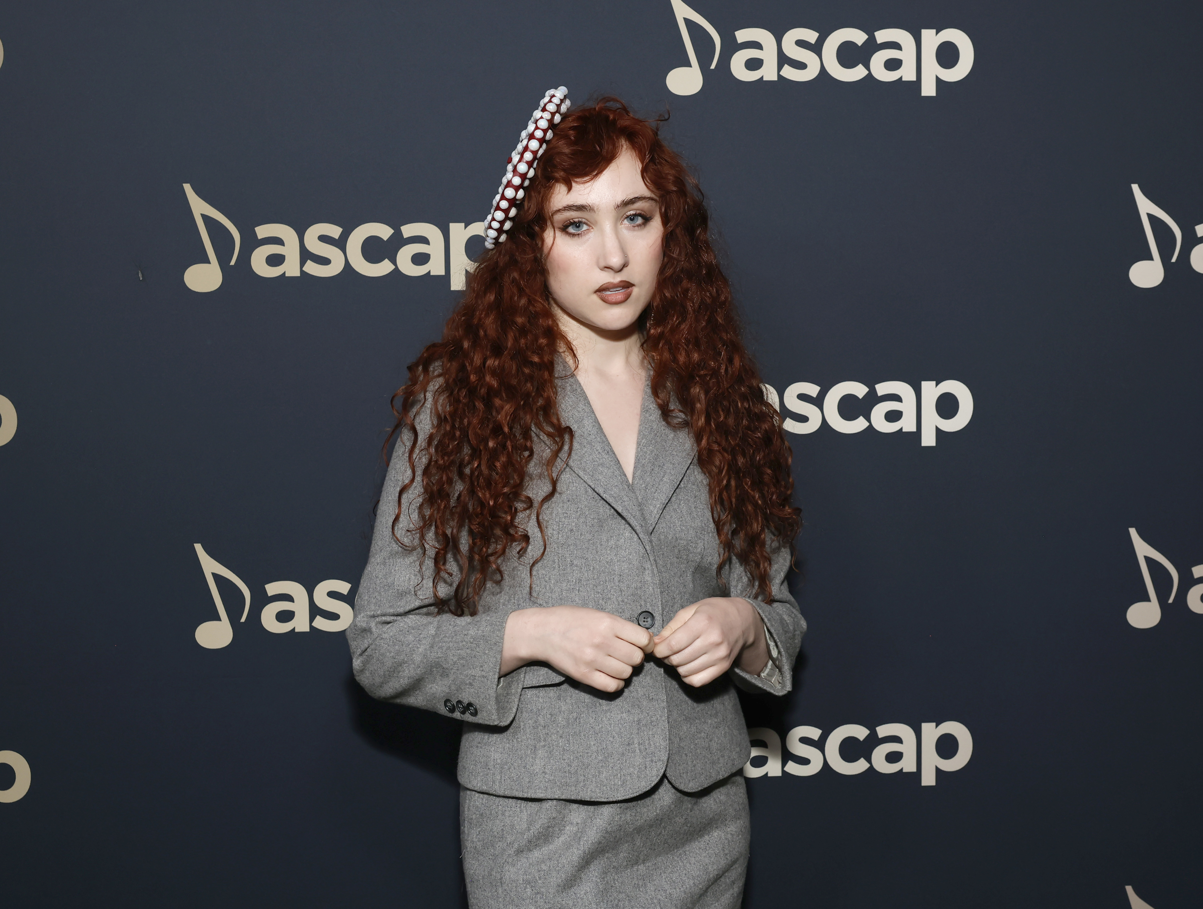 A woman with long red curly hair, wearing a gray suit and a decorative headband, poses in front of an "ascap" backdrop at a celebrity event