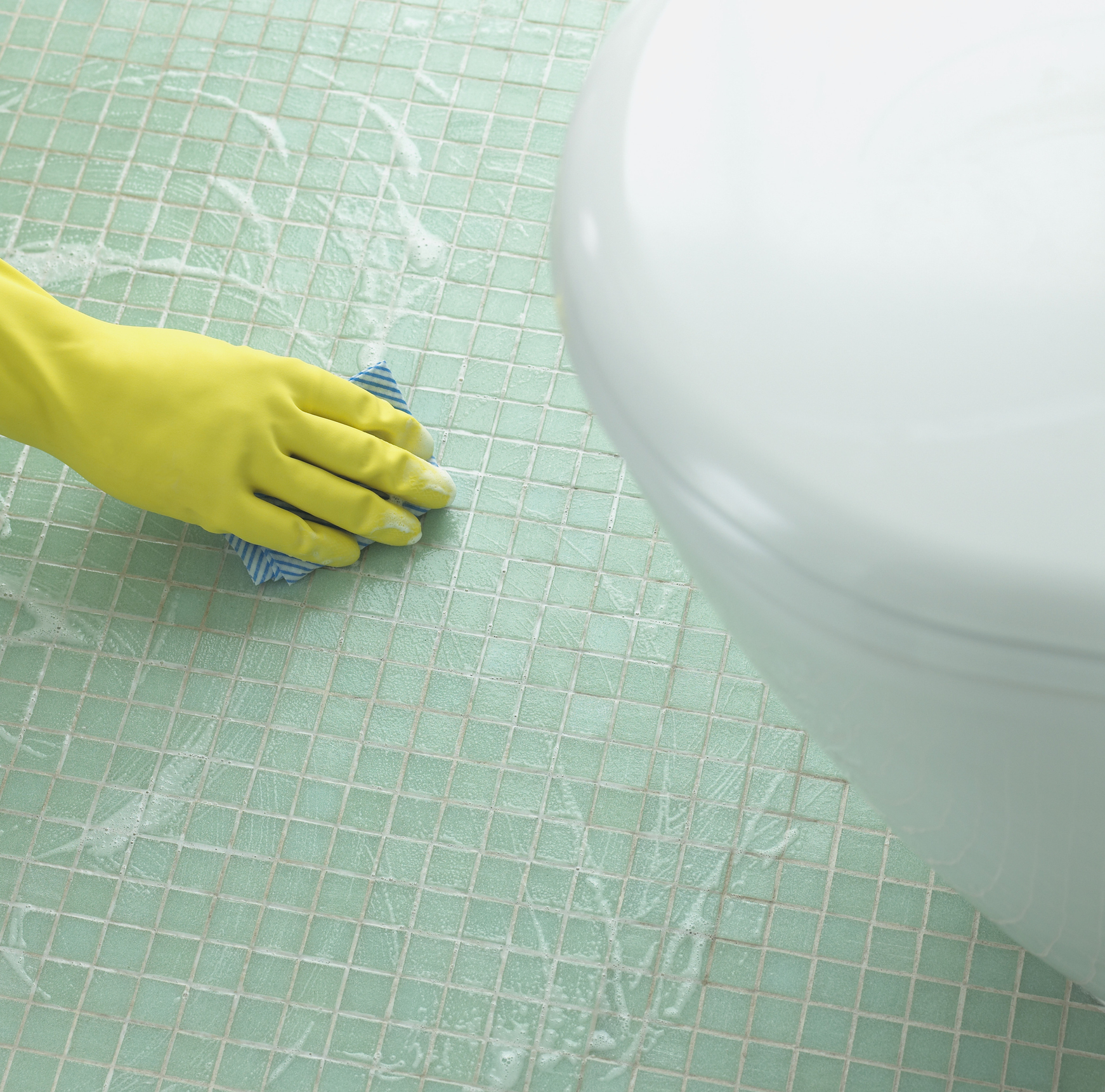 A hand wearing a yellow glove scrubs a tiled floor near a white toilet with a blue and white cloth