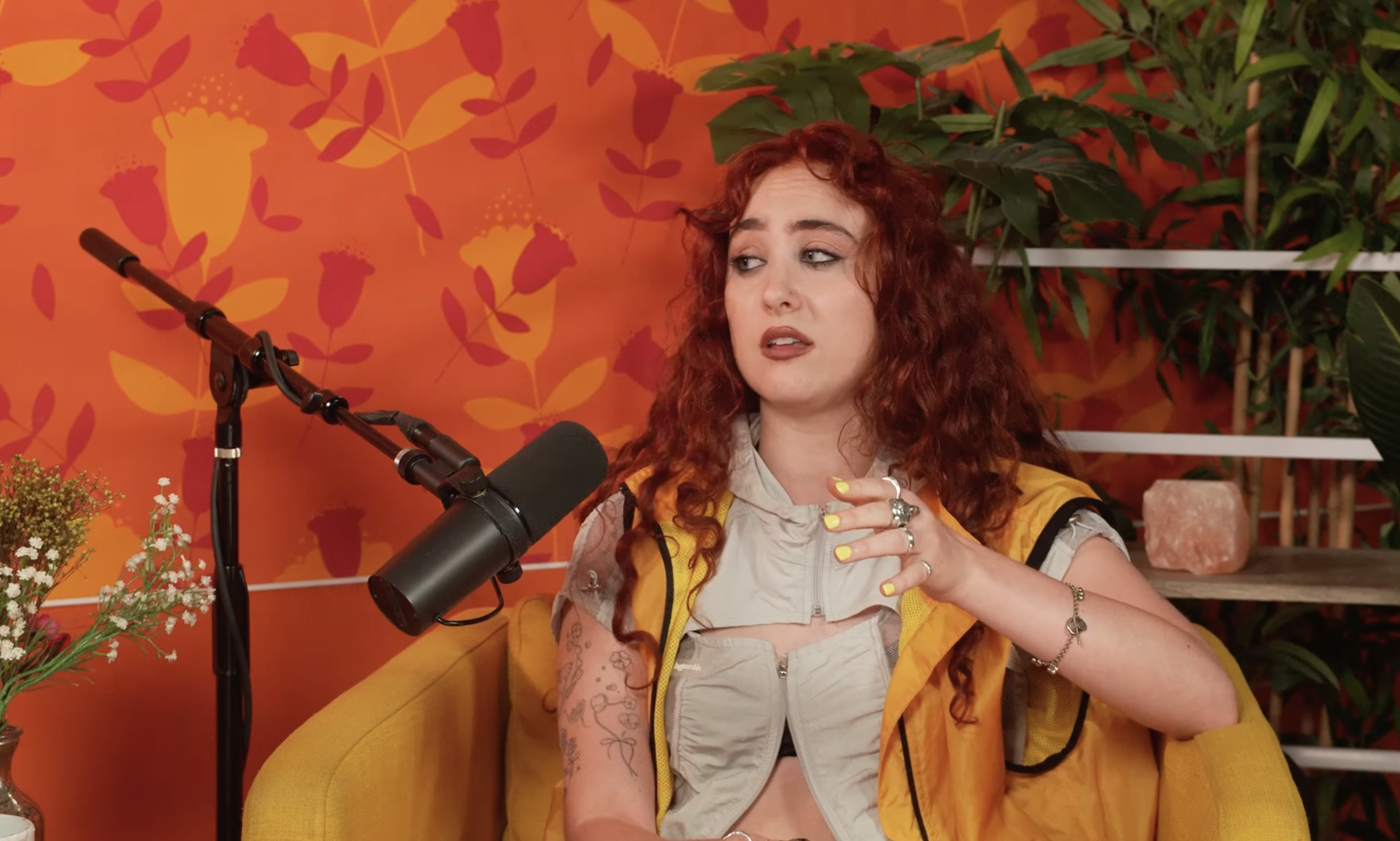 A woman with curly red hair sits on a yellow chair, speaking into a microphone. She wears a light-colored top with a yellow vest and has tattoos on her arms