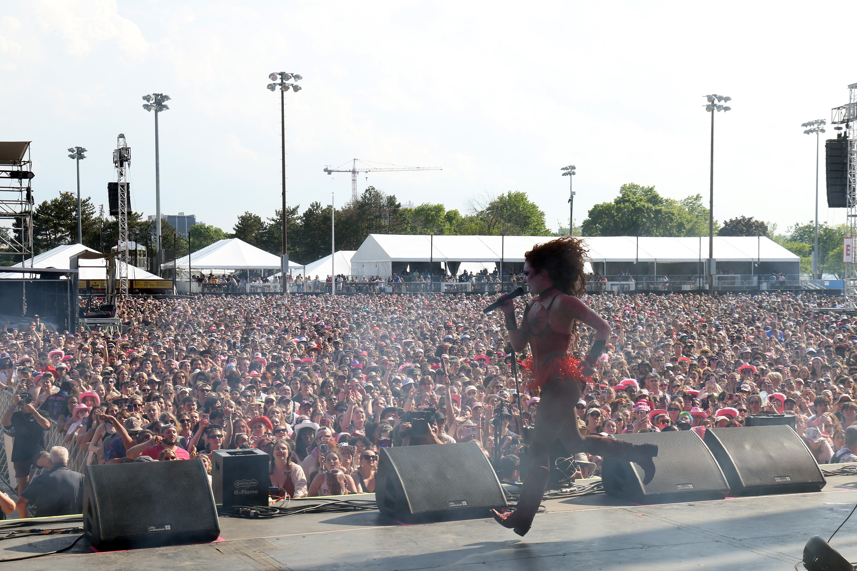 A performer dressed in a red outfit is singing on stage at a large outdoor music festival, with a huge crowd in the background