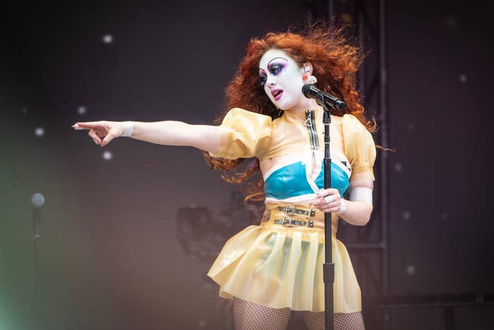 A singer in a theatrical outfit, including a sheer puff-sleeve top and a short skirt, energetically performing on stage while pointing
