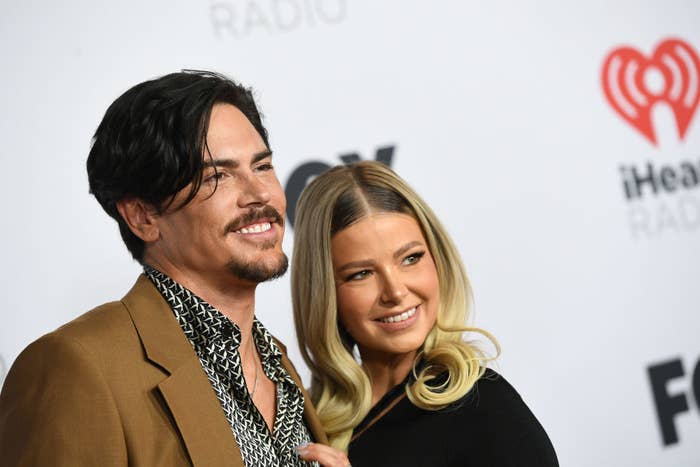 Tom Sandoval and Ariana Madix smile together at the iHeartRadio event, with Tom wearing a brown jacket and patterned shirt, and Ariana in a black outfit