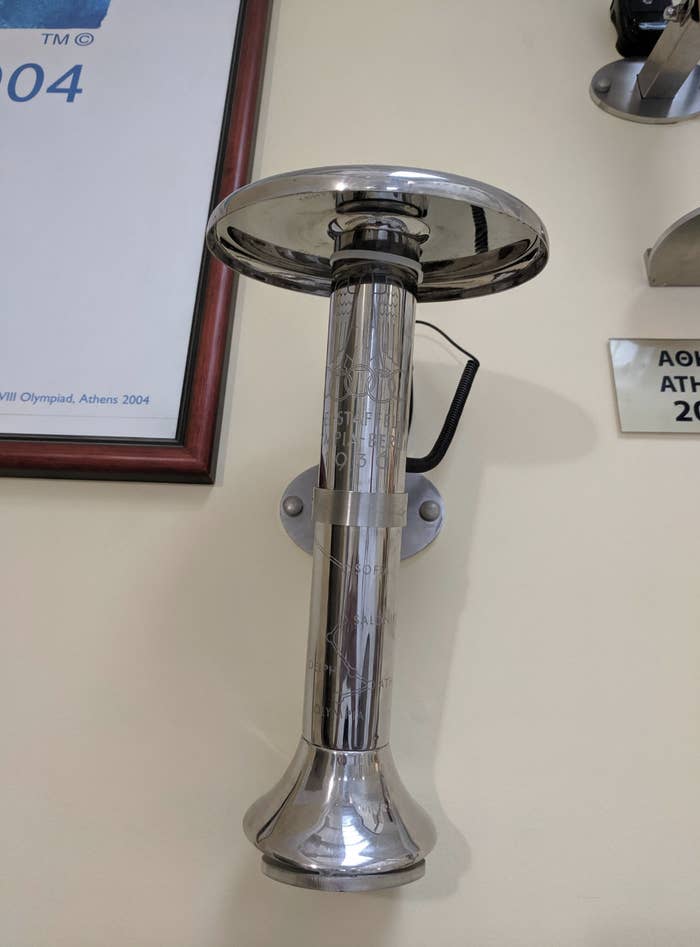 A metallic Olympic torch mounted on a wall with a small engraved plaque beside it. It is part of a displayed collection