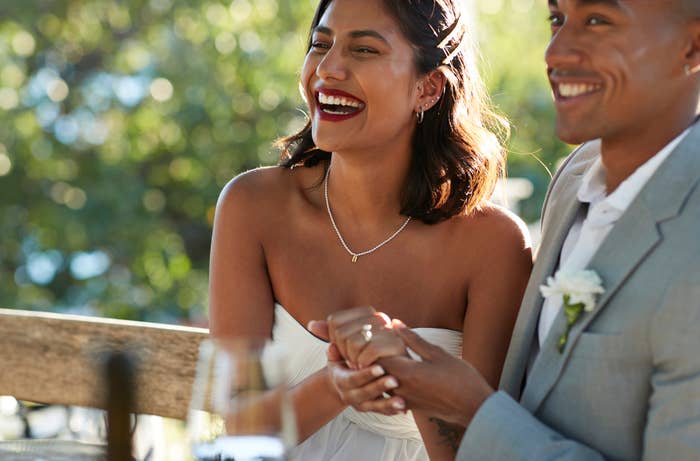 A woman in a strapless dress and a man in a suit sit outdoors, smiling and holding hands, possibly during a celebratory event