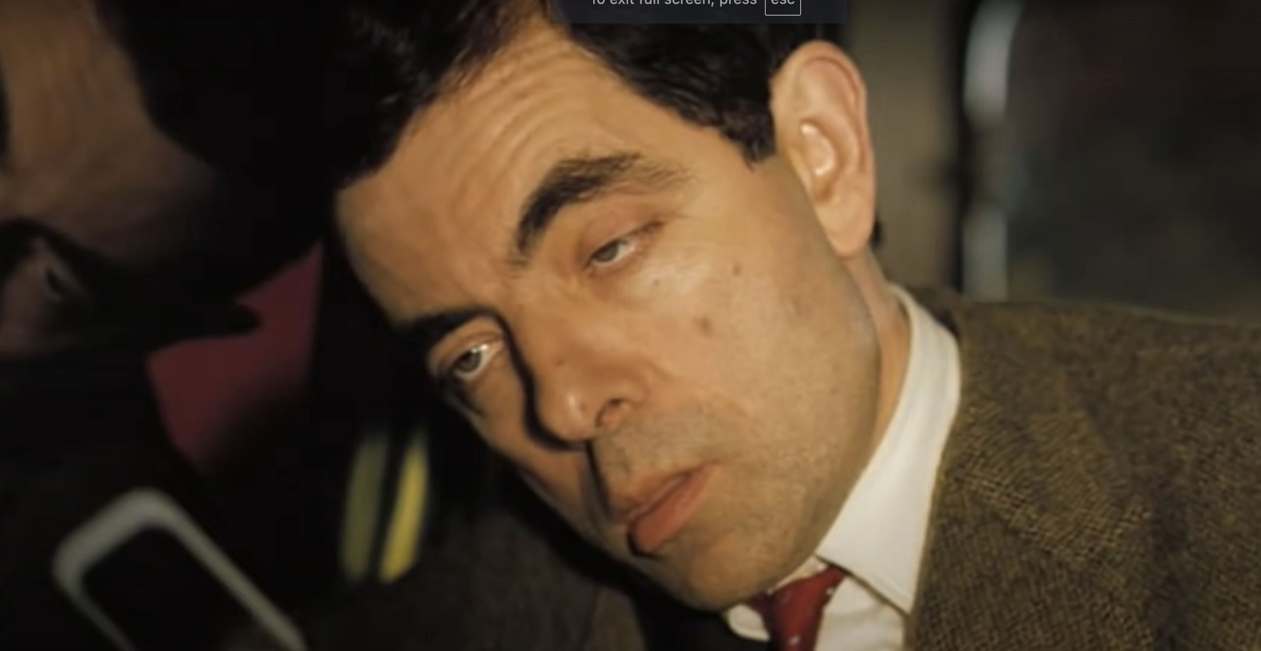 Rowan Atkinson, in character as Mr. Bean, appears tired with droopy eyes and a disheveled suit and tie during a close-up shot