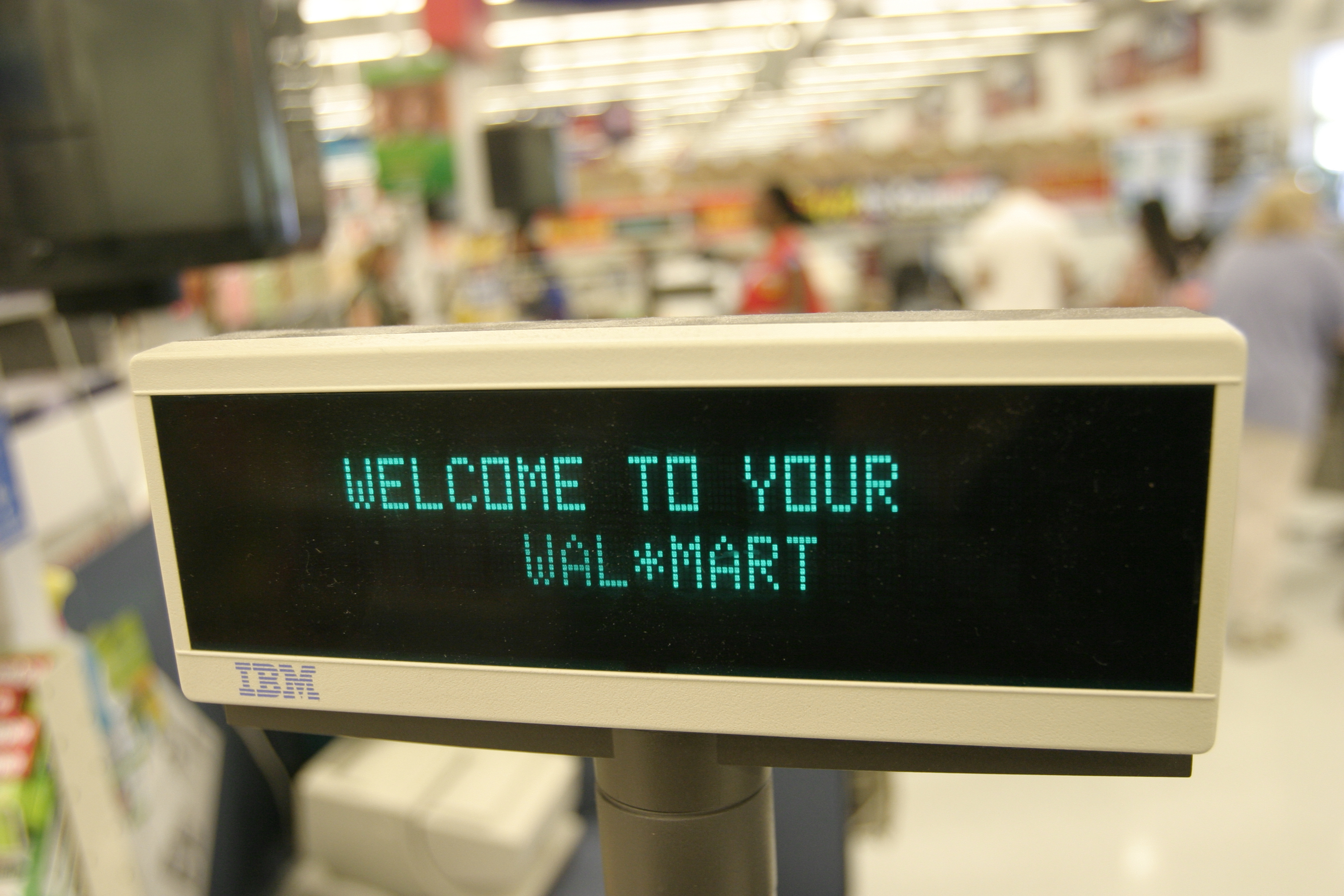 A digital display at the checkout of a Walmart store reads &quot;WELCOME TO YOUR WAL*MART&quot; in green text. The background is blurred, showing a store setting