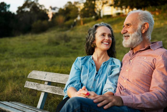 Older couple sitting on a park bench, smiling warmly at each other. The woman has long curly hair; the man has a beard