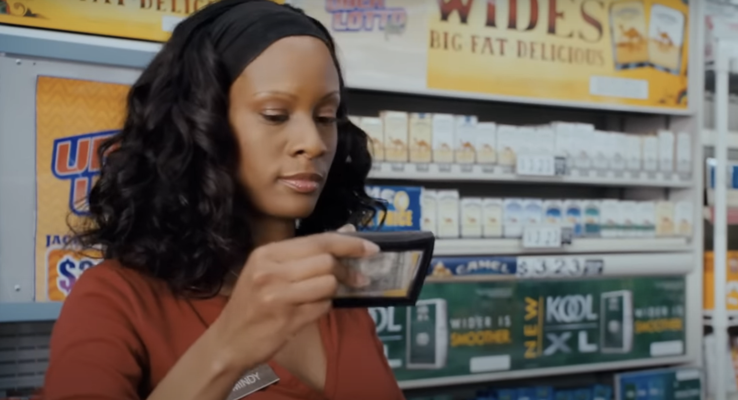 A person with black wavy hair, wearing a name tag and a red shirt, holds a wallet in a store aisle stocked with cigarette packs