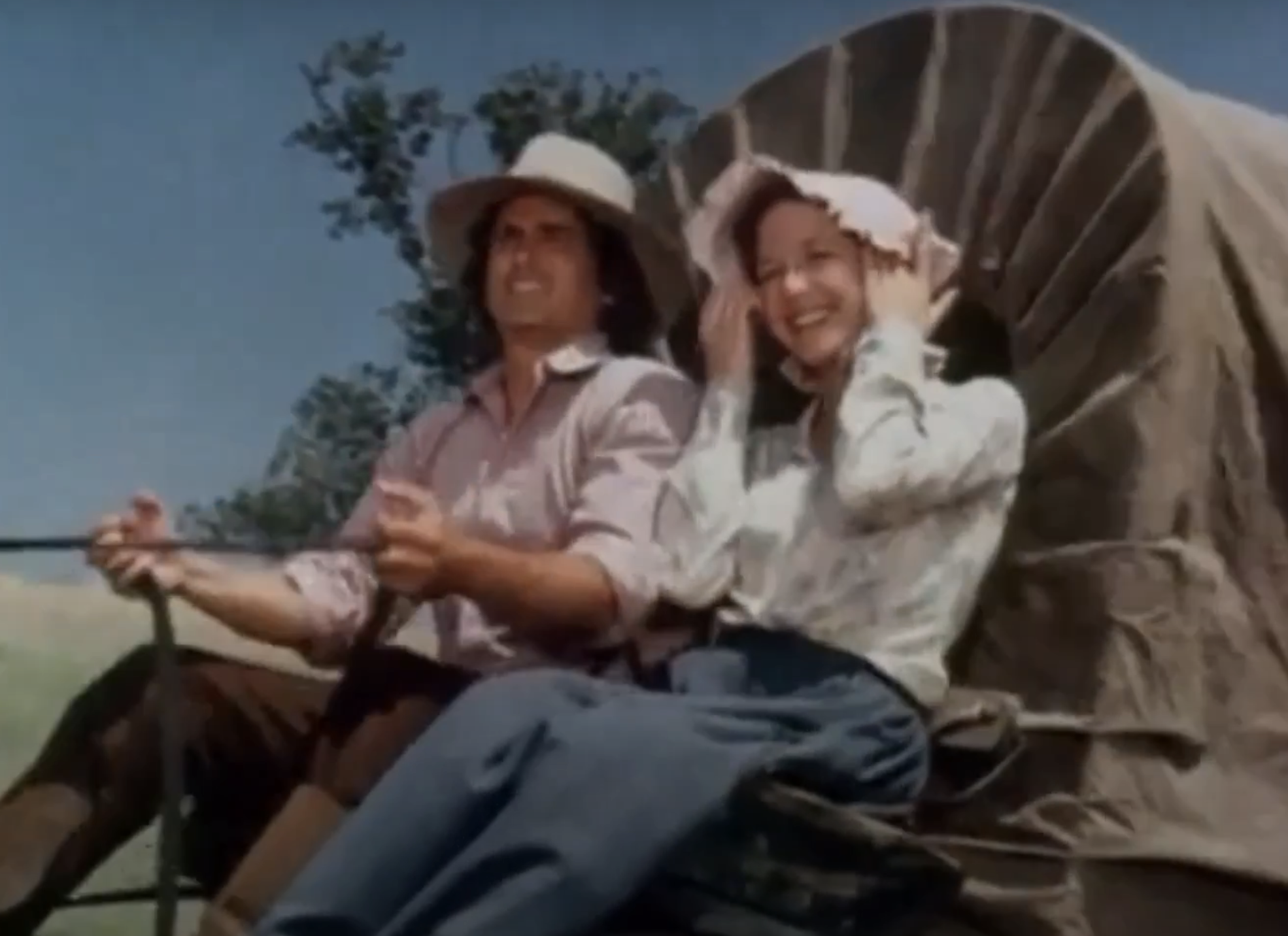 Michael Landon and Karen Grassle sit on a horse-drawn wagon in a vintage countryside setting. Landon holds the reins, while Grassle adjusts her bonnet, smiling