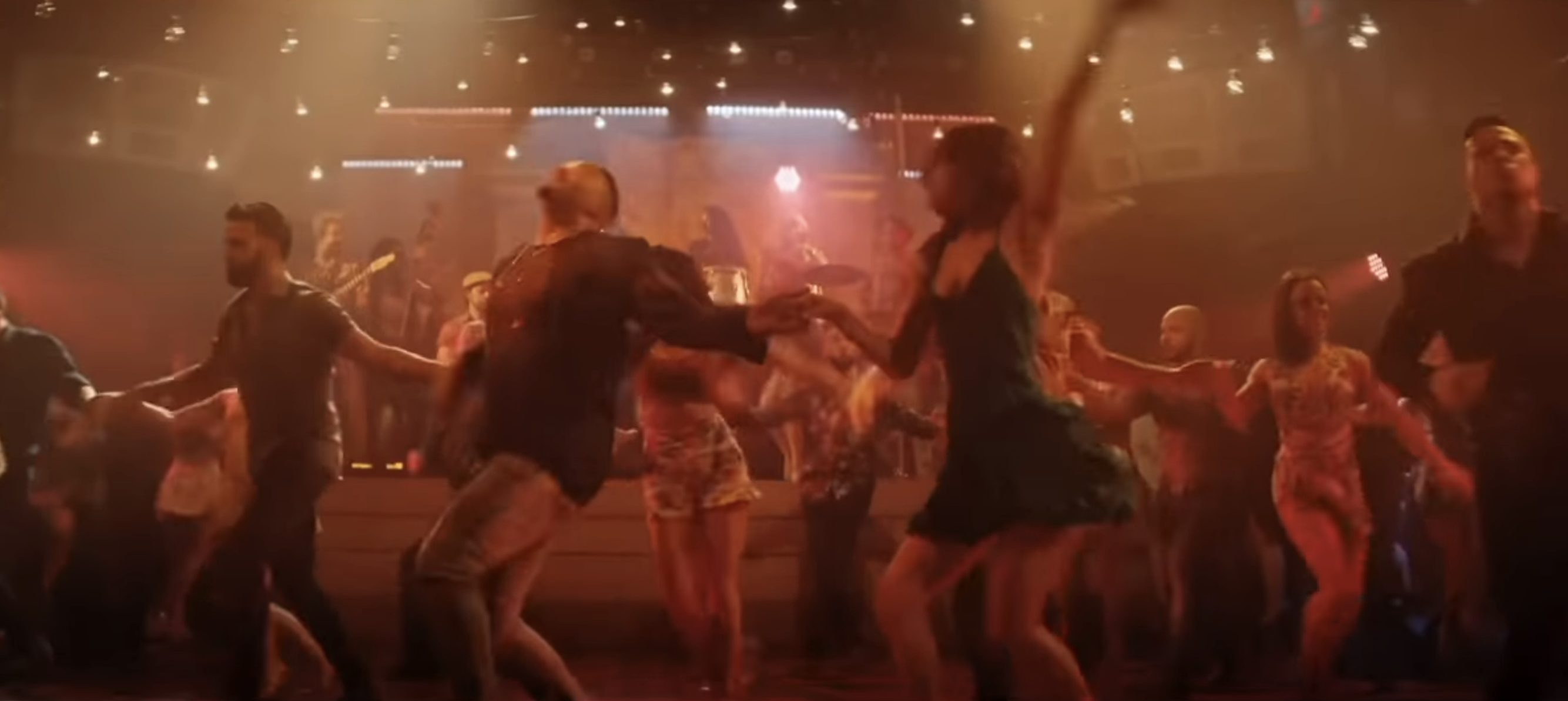 Group of people dancing energetically in a lively, dimly lit dance club with a band playing on stage in the background