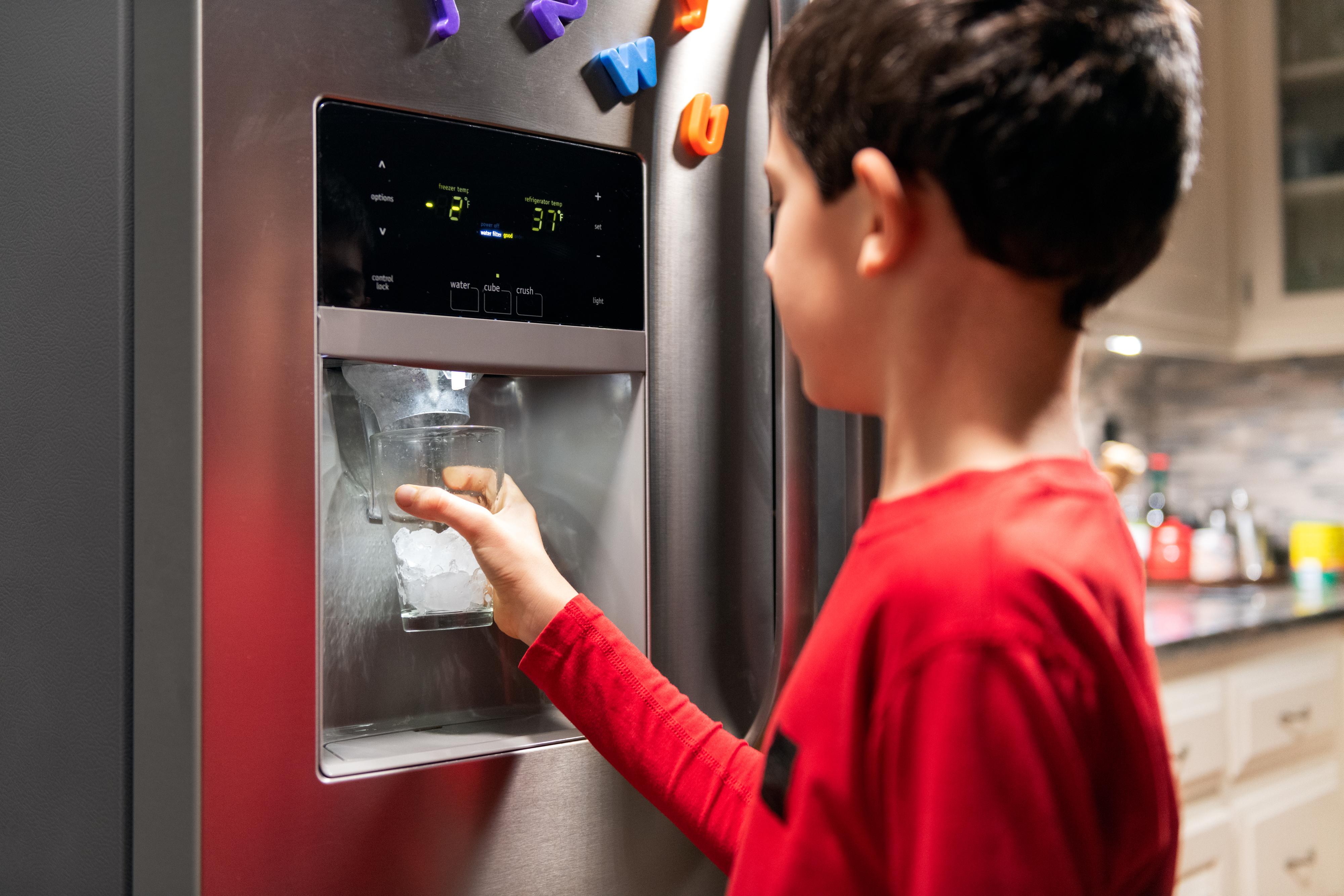 A young boy in a red shirt fills a glass with ice from a refrigerator dispenser in a kitchen