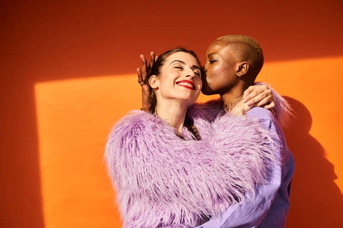 Two people embrace happily against an orange background. One wears a fluffy jacket, and the other gives a kiss on the cheek. Names unknown