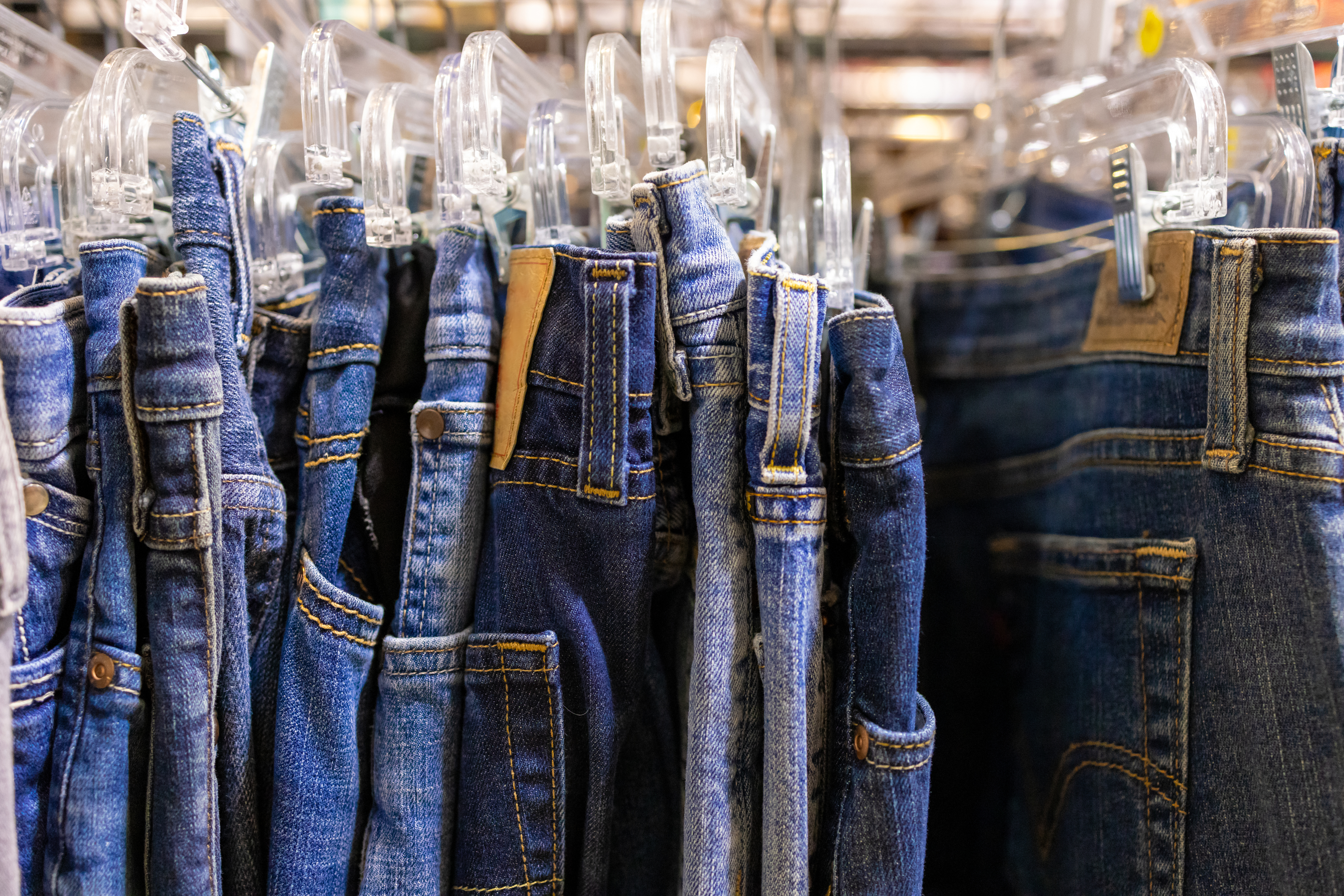 Multiple pairs of blue jeans hanging on clear plastic hangers in a clothing store. Some jeans have visible pockets and belt loops