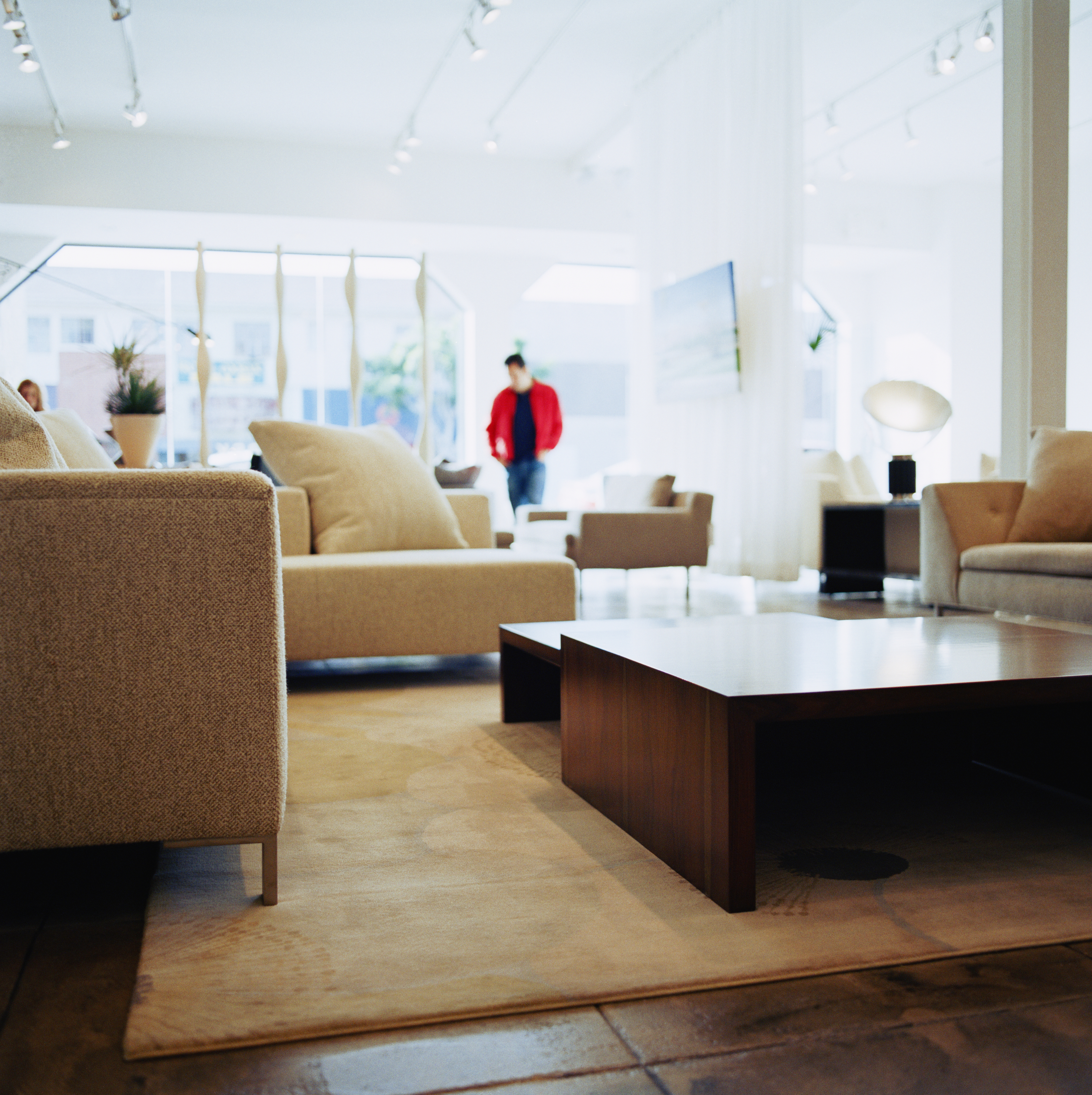 Modern living room with beige furniture, a wooden coffee table, and large windows. Two people in casual clothing are standing in the background