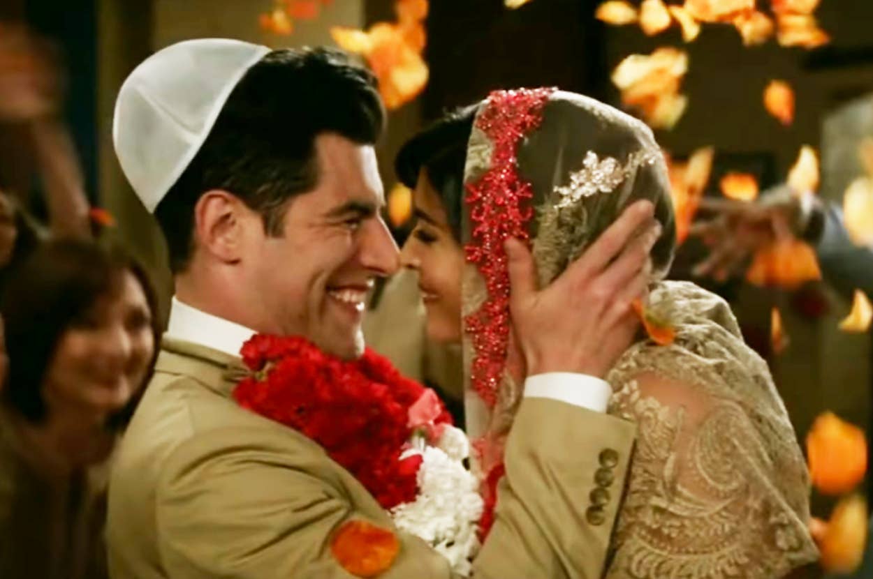 Max Greenfield and Hannah Simone smile and embrace at their cultural wedding ceremony, surrounded by flower petals