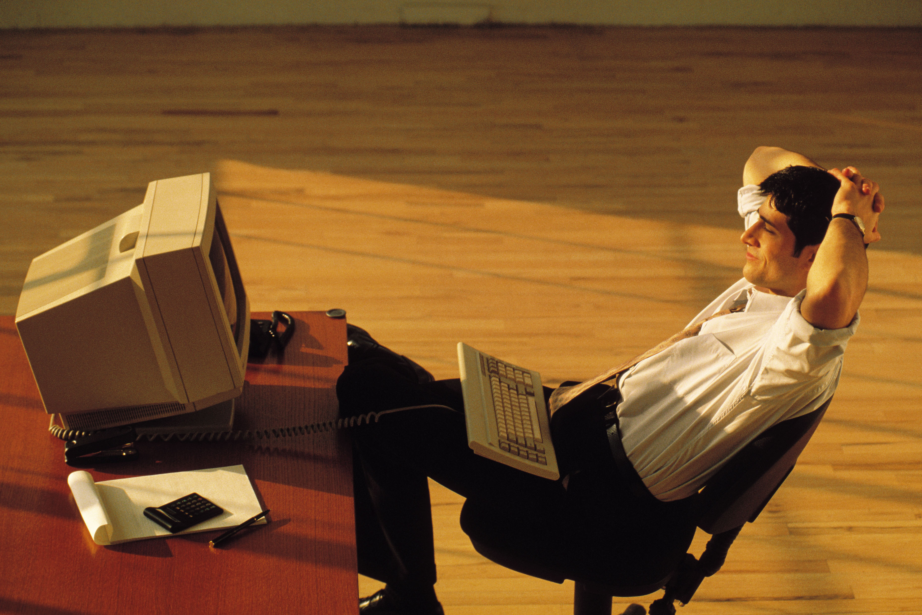 A man in a dress shirt and tie leans back in his chair, with his hands behind his head, in front of a computer on a large wooden desk