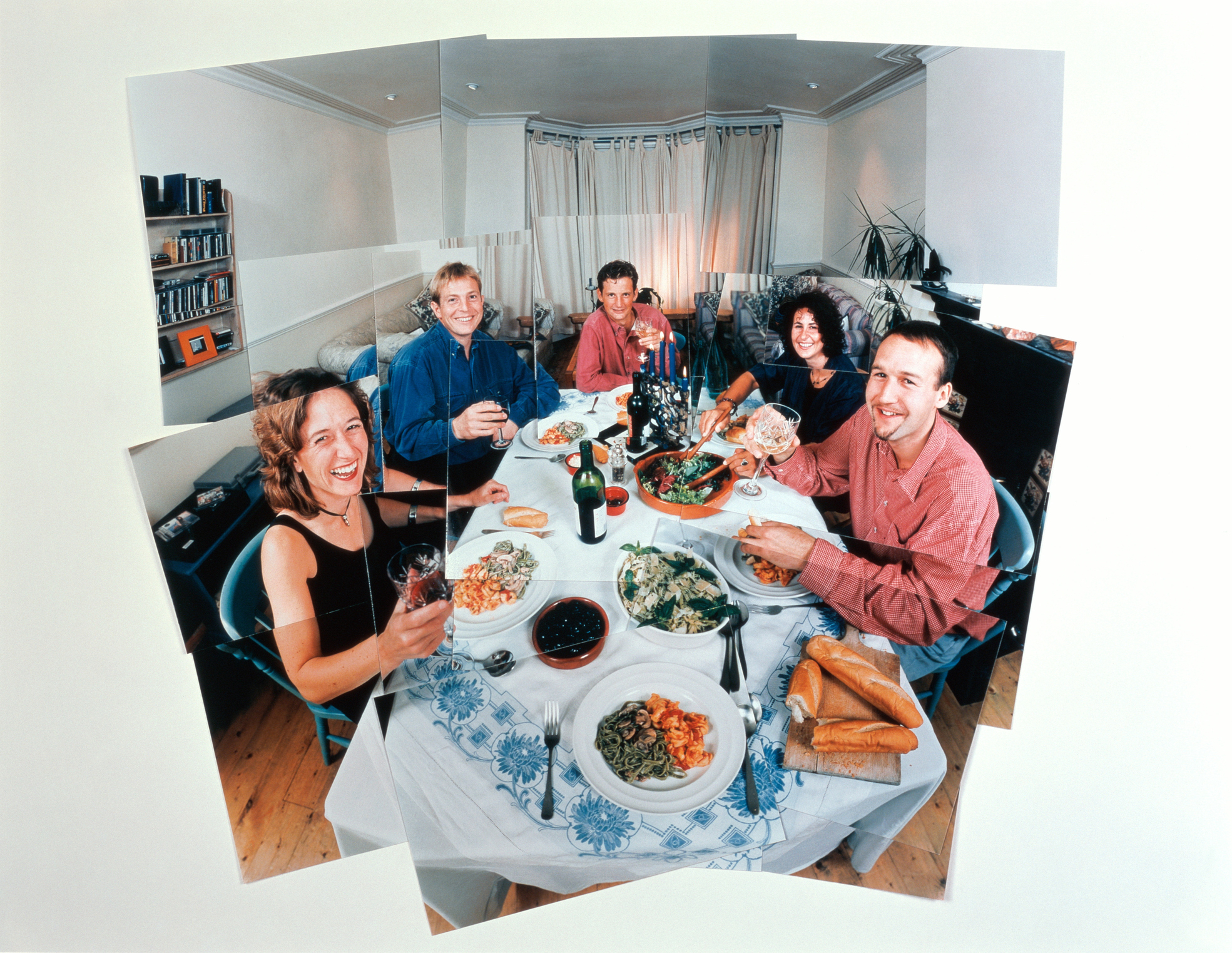 People seated around a dining table enjoying a meal together, smiling and raising glasses. The table is set with various dishes, candles, and bread