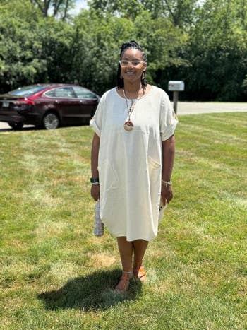 A reviewer stands connected writer successful a loose-fitting light-colored dress, sandals, and accessories. She holds a tiny clutch and smiles astatine the camera