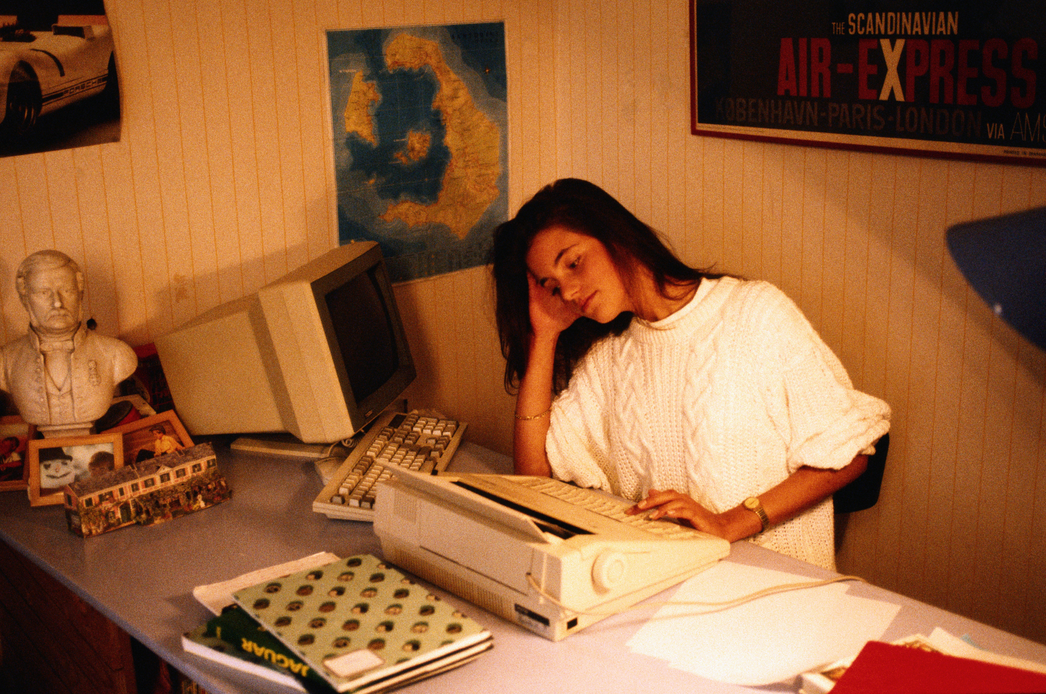 A woman in a knitted sweater sits at a desk with her head resting on her hand, surrounded by retro office equipment including a typewriter and an old computer