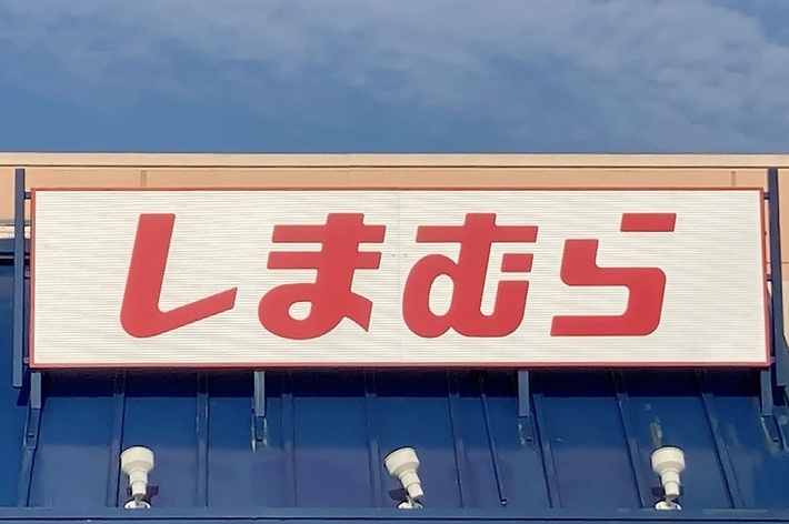 Sign in Japanese text that reads “しまむら” (Shimamura) above the entrance of a store, with a blue sky in the background