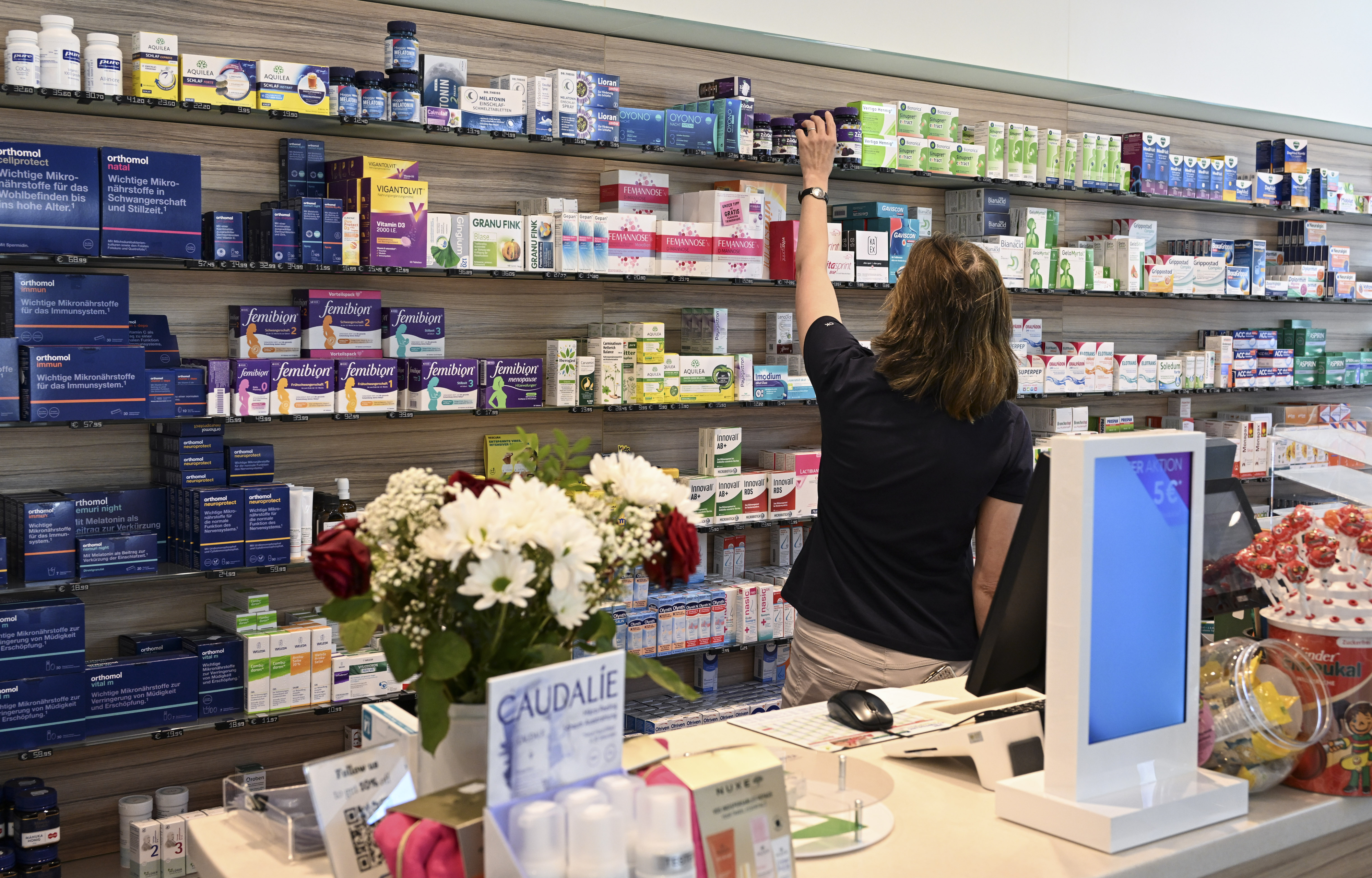 A person reaches for a product on a shelf in a well-stocked pharmacy with various health items, including a display of flowers on the counter