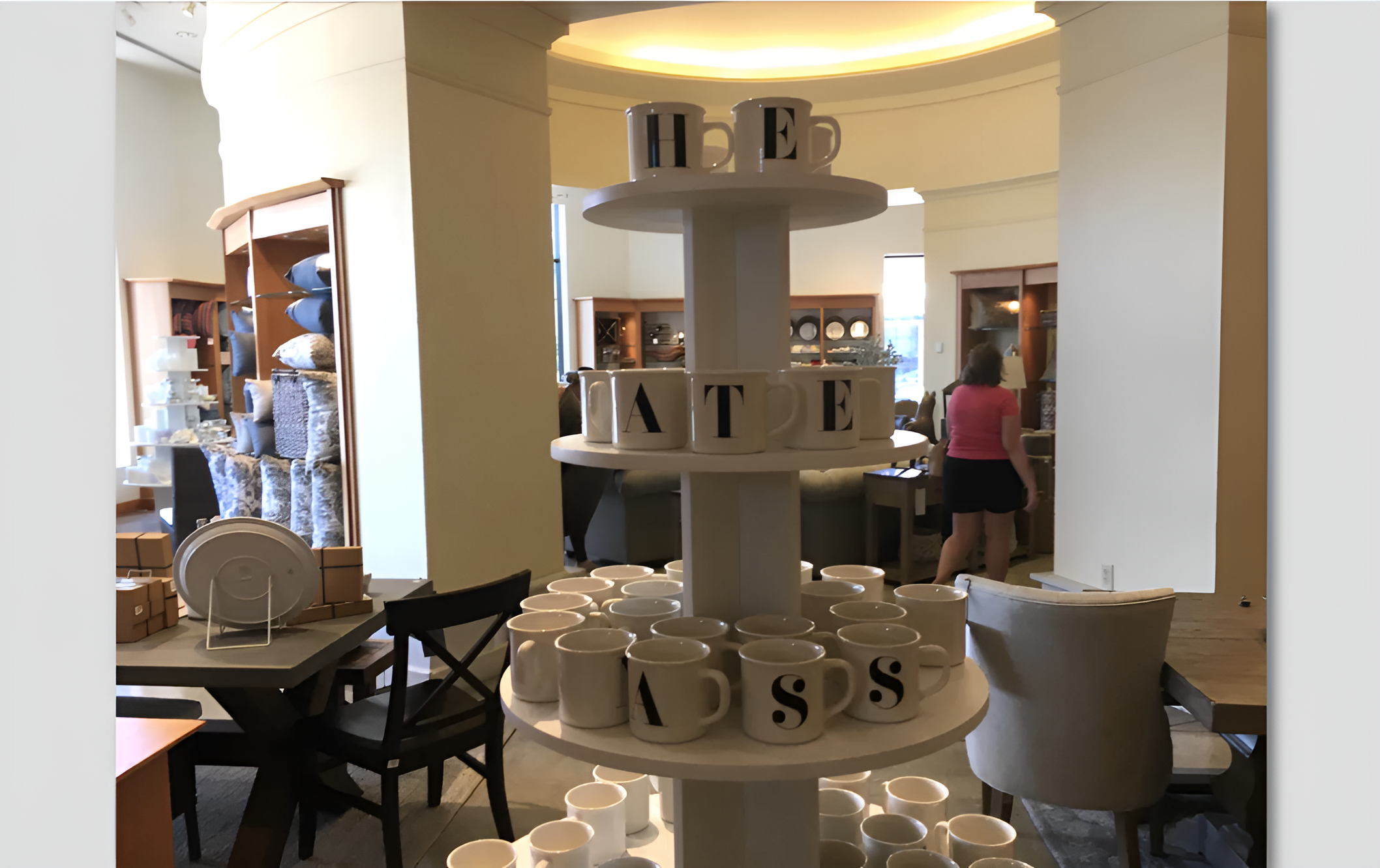 Mugs on a display stand spell out "HE ATE ASS." The image is taken inside a store with shelves and people visible in the background