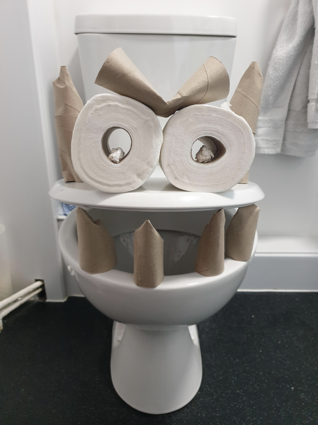 Toilet humorously decorated with empty toilet paper rolls to appear like a face, featuring a toothy grin and eyes
