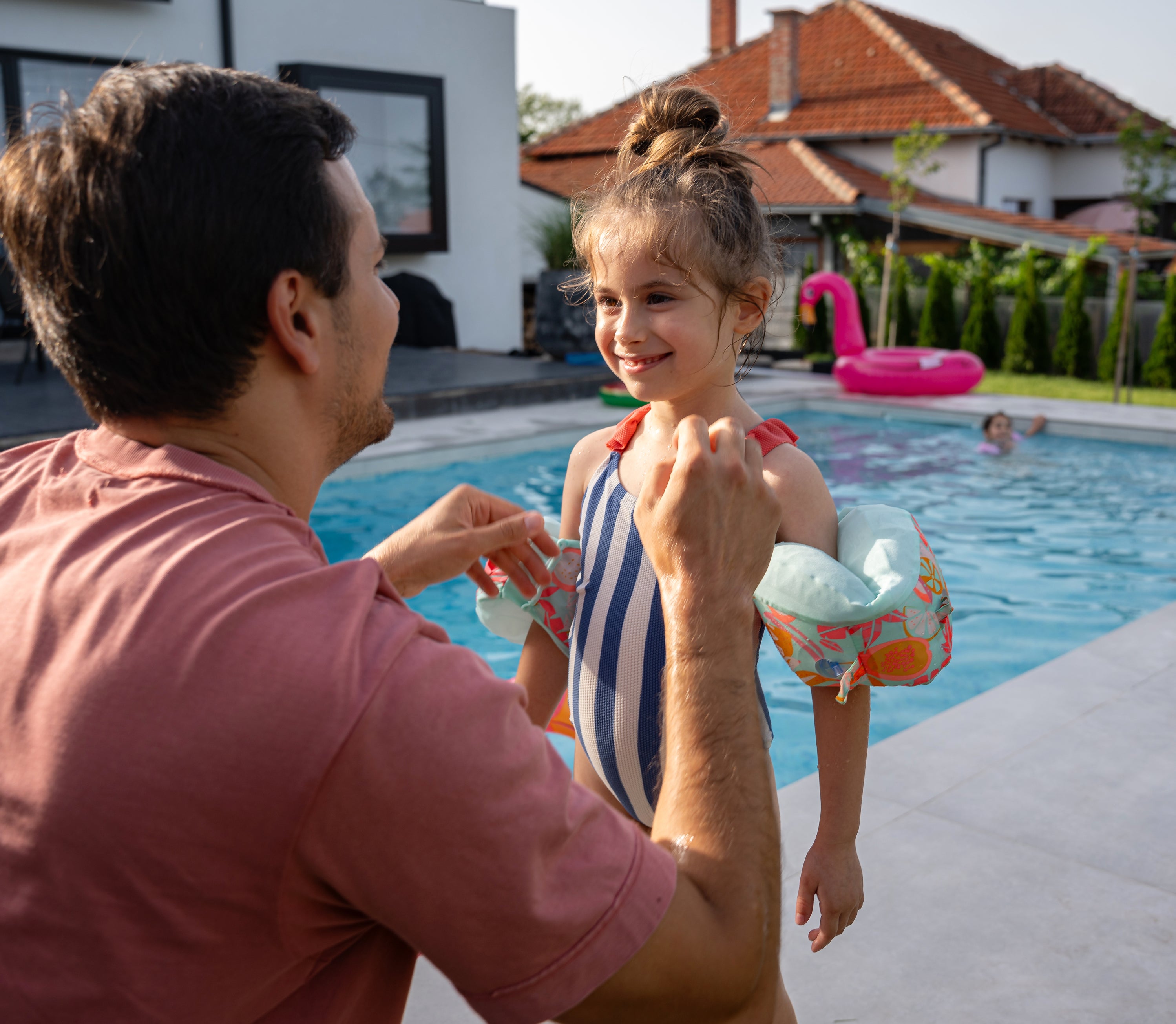 A man and a young girl in a swimsuit with arm floats smile near a pool at a suburban home. Another child is swimming in the background