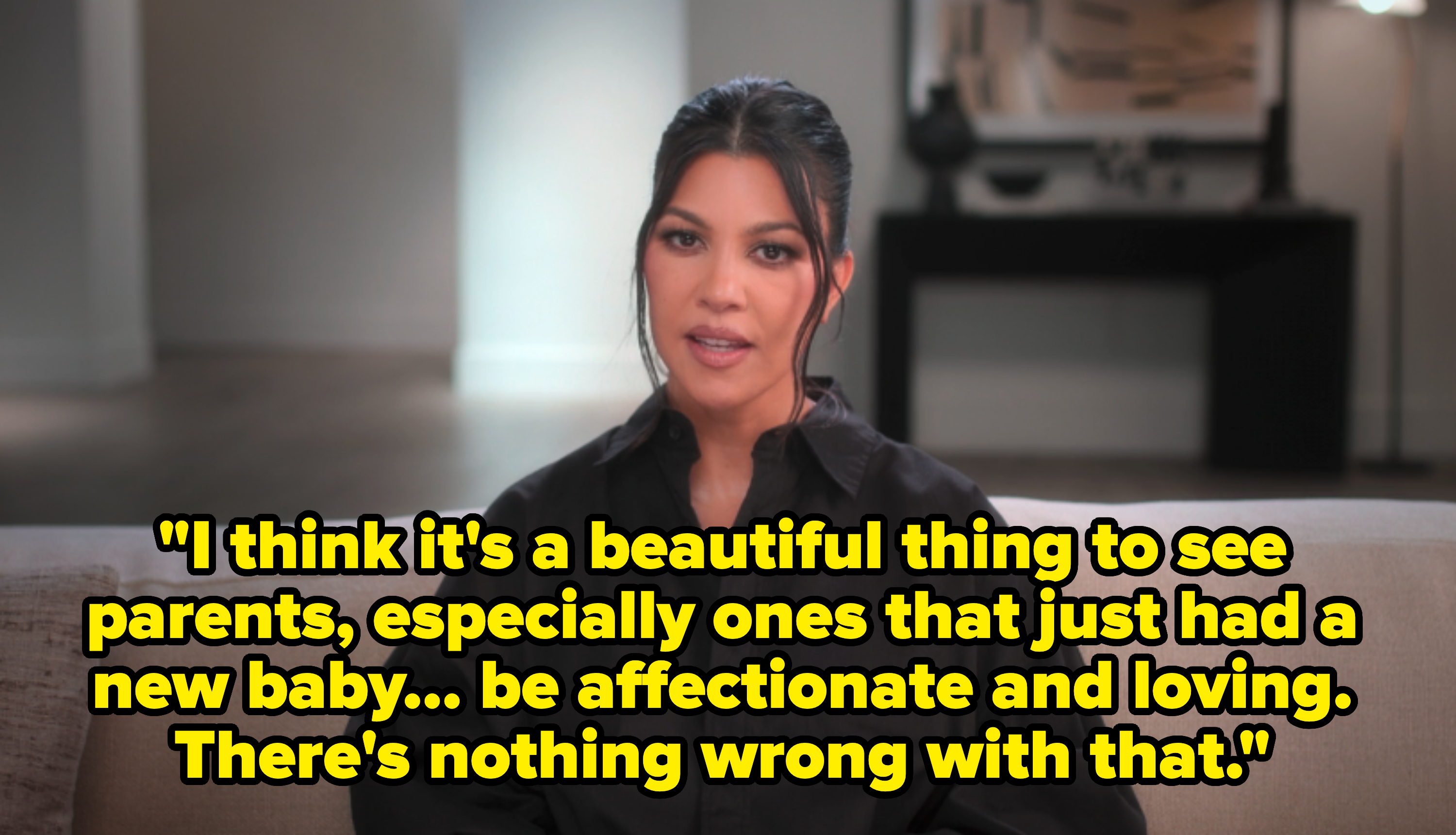 Kourtney Kardashian sits on a beige sofa, wearing a loose-fitting, dark button-up shirt, speaking directly at the camera in an indoor setting