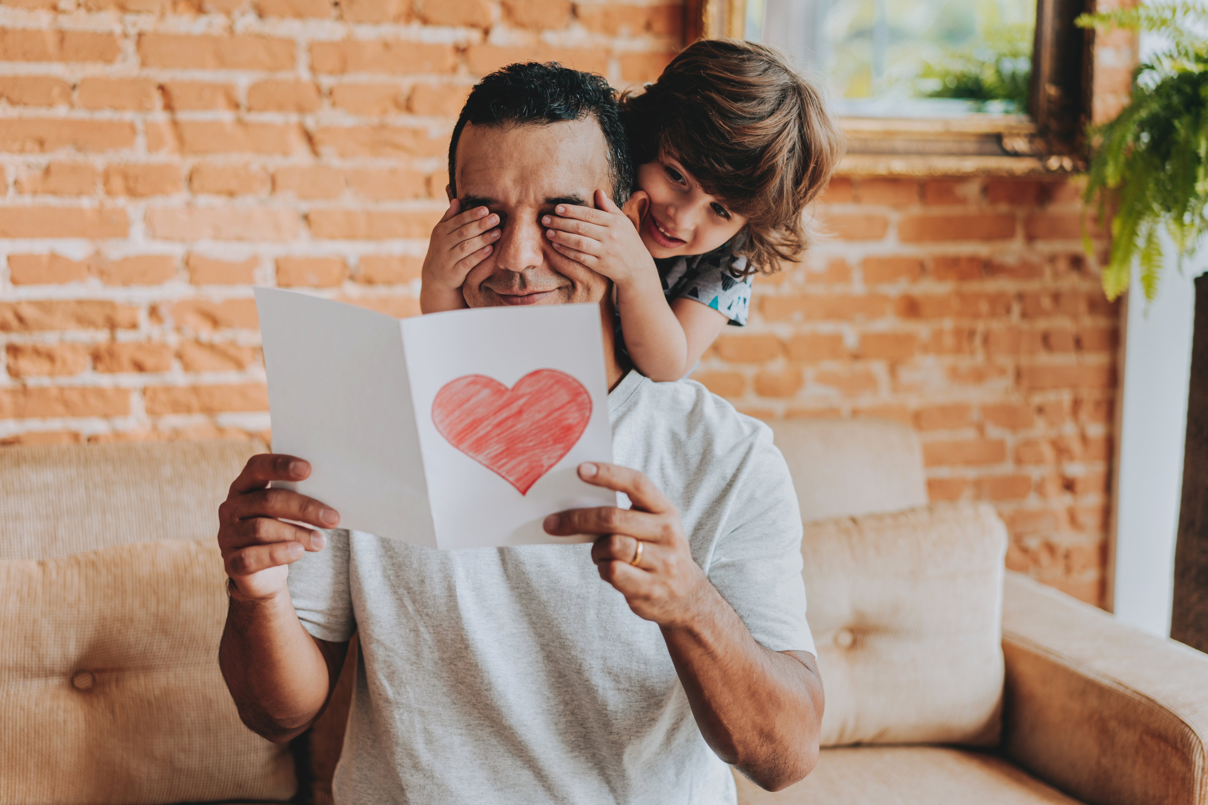 A man sits on a couch reading a card with a red heart on it, as a child playfully covers his eyes from behind