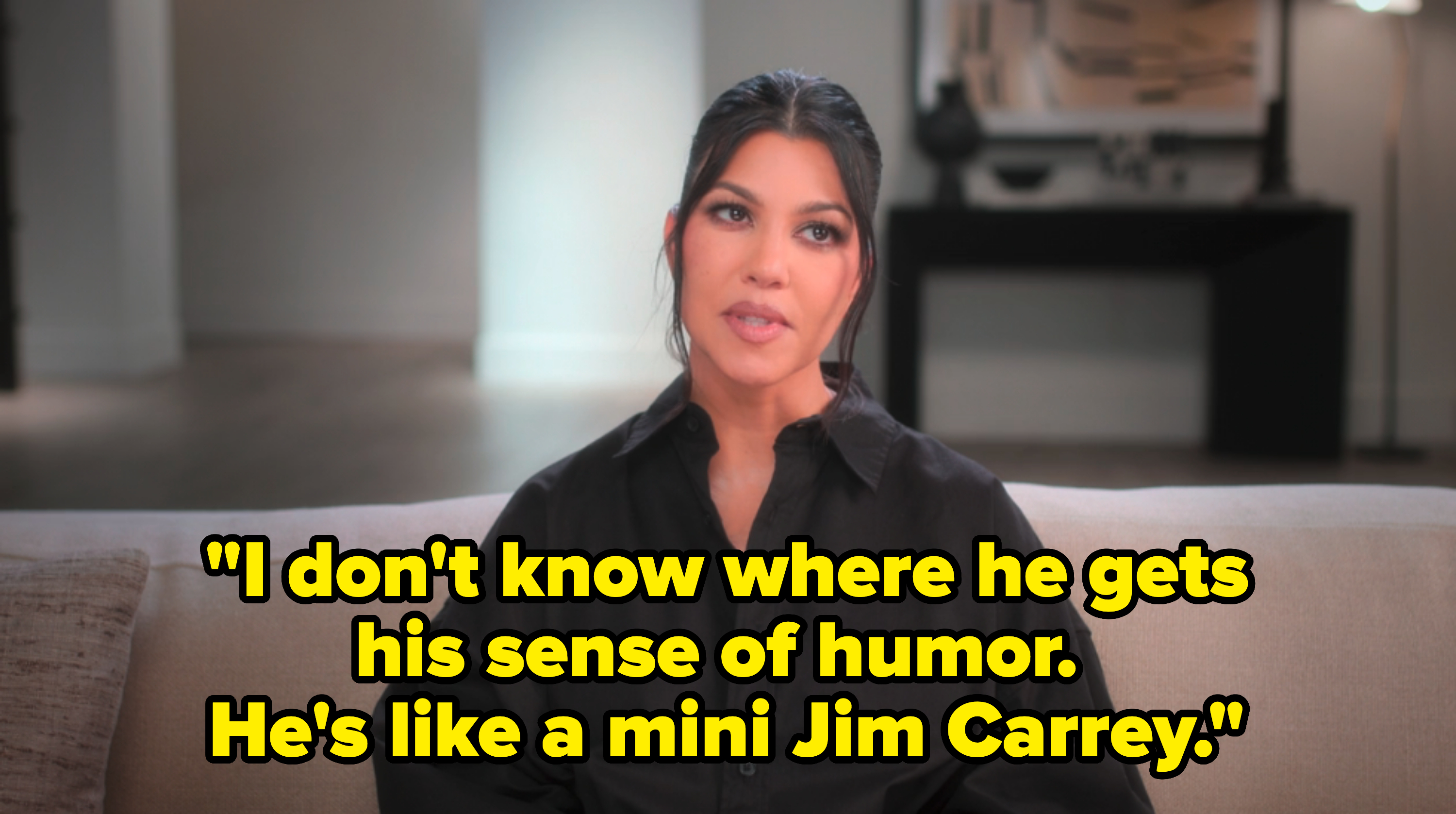 Kourtney Kardashian sitting on a couch, wearing a black button-up shirt, in a modern living space