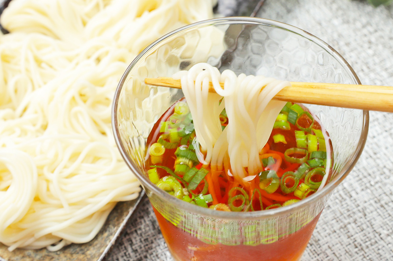 そうめんが入ったガラスの器に箸で麺を持ち上げるシーン。背景に皿に盛られたそうめんが見える。