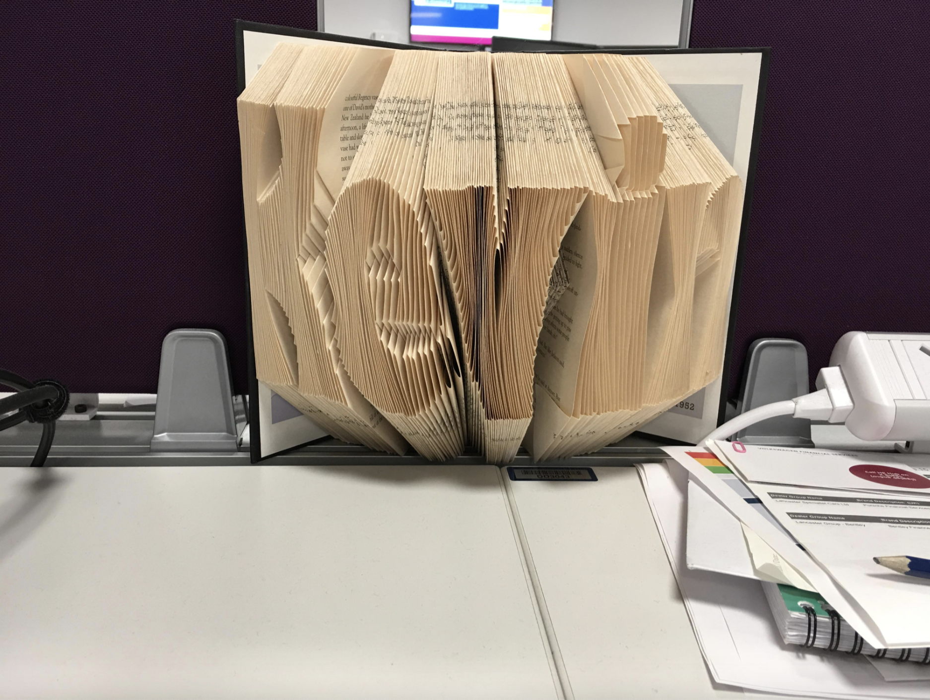 An opened book with pages folded to spell out "Kevin," placed on a desk with various papers and office supplies