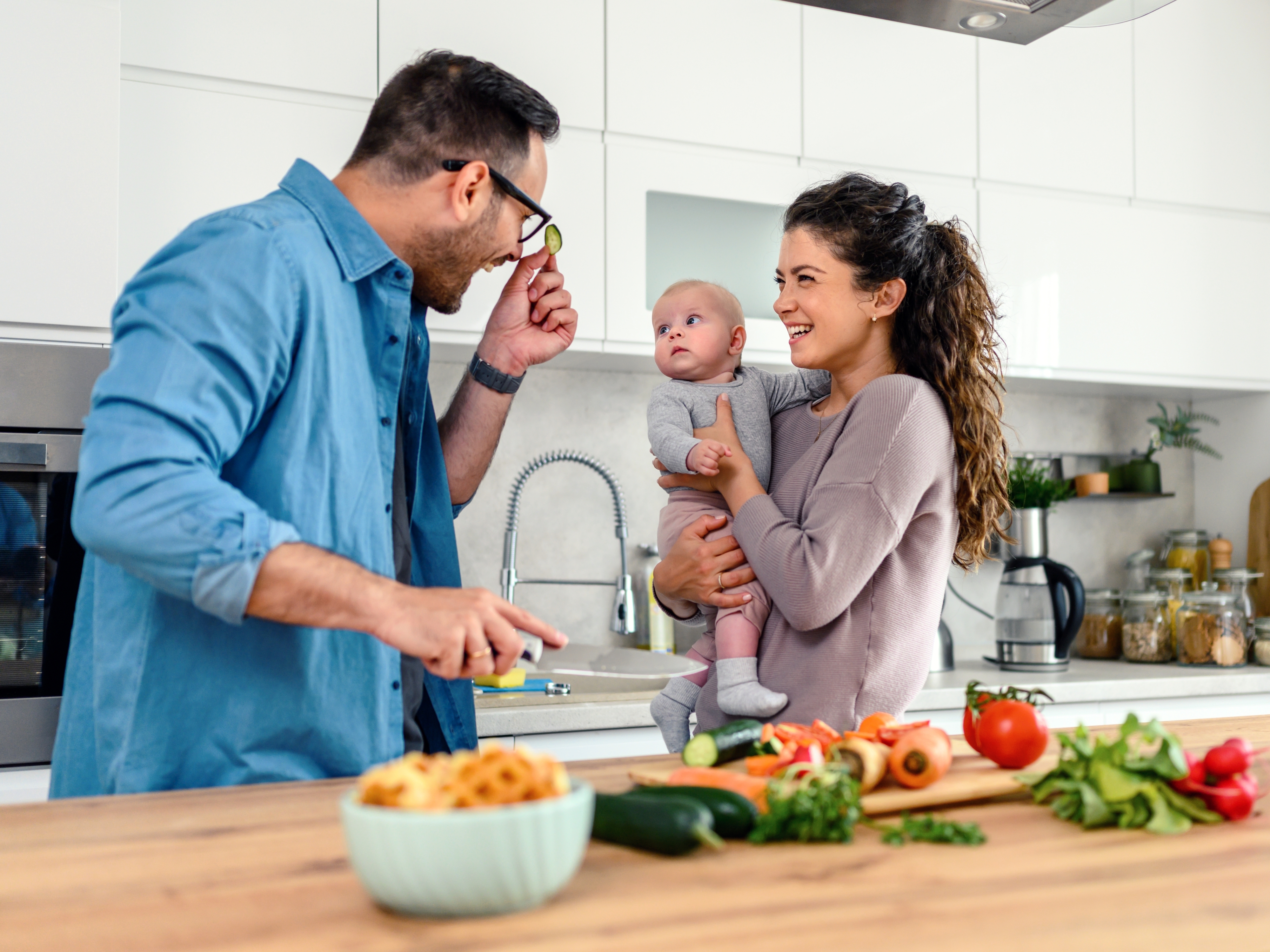 A man playfully adjusts his glasses, making a baby he is holding laugh, while a woman smiles at them. They are in a kitchen with various vegetables on the countertop
